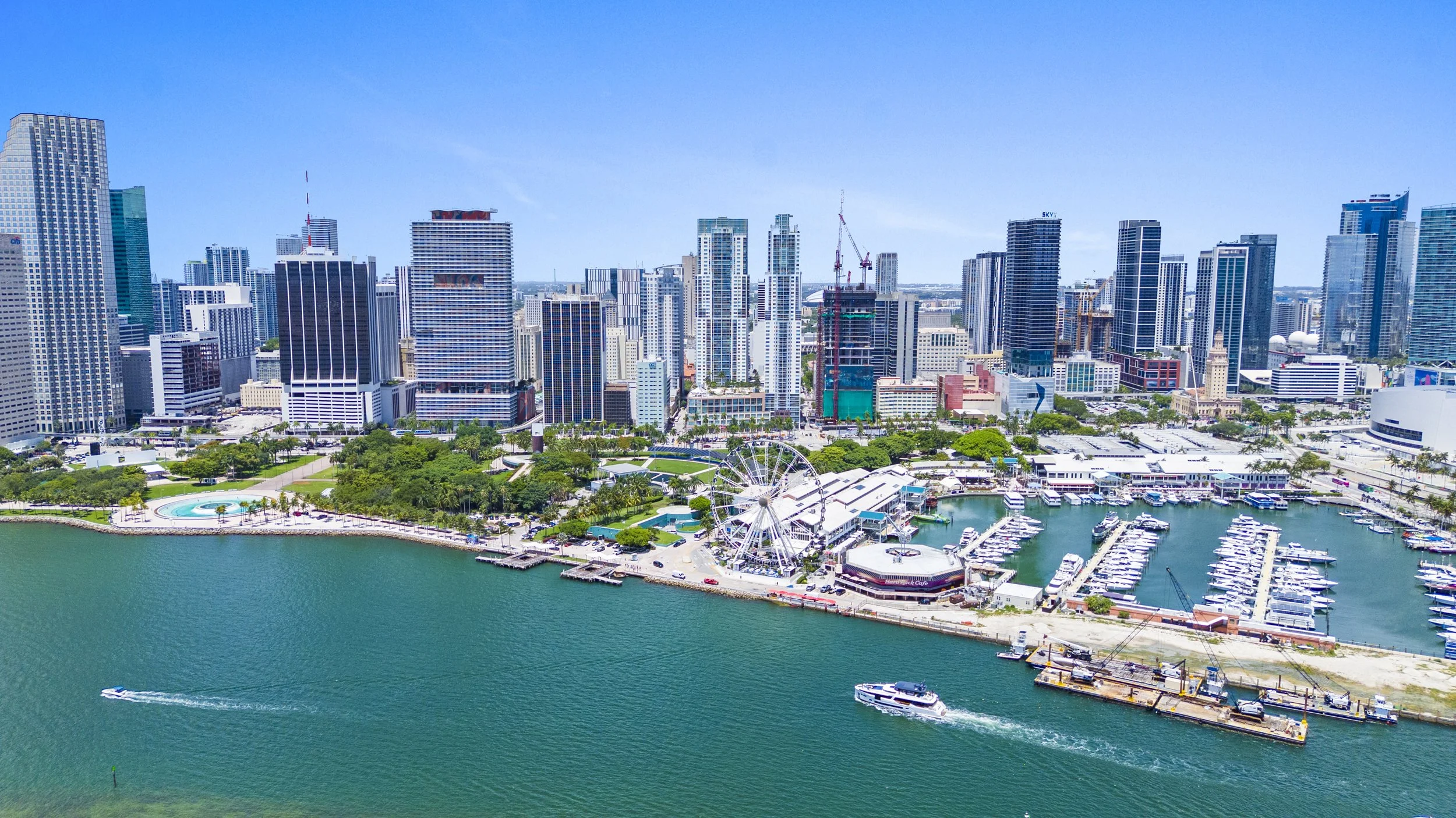 Aerial view of a city skyline with tall modern skyscrapers, a waterfront with boats docked, a Ferris wheel, and green parks under a clear blue sky.