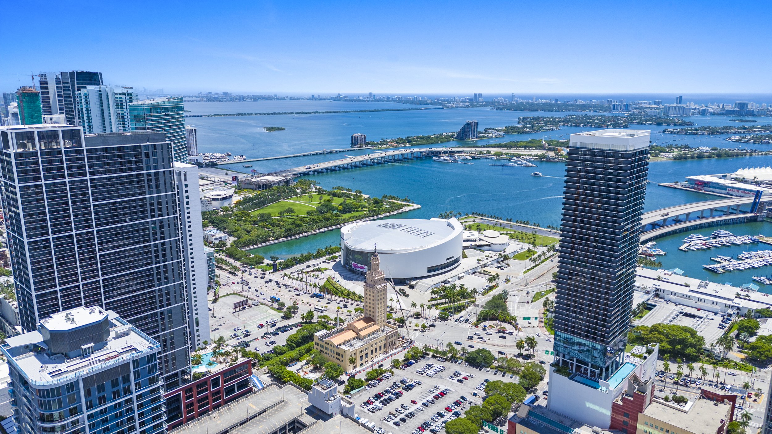 Aerial view of Miami skyline with high-rise buildings, waterways, a park, and marina on a bright, clear day.
