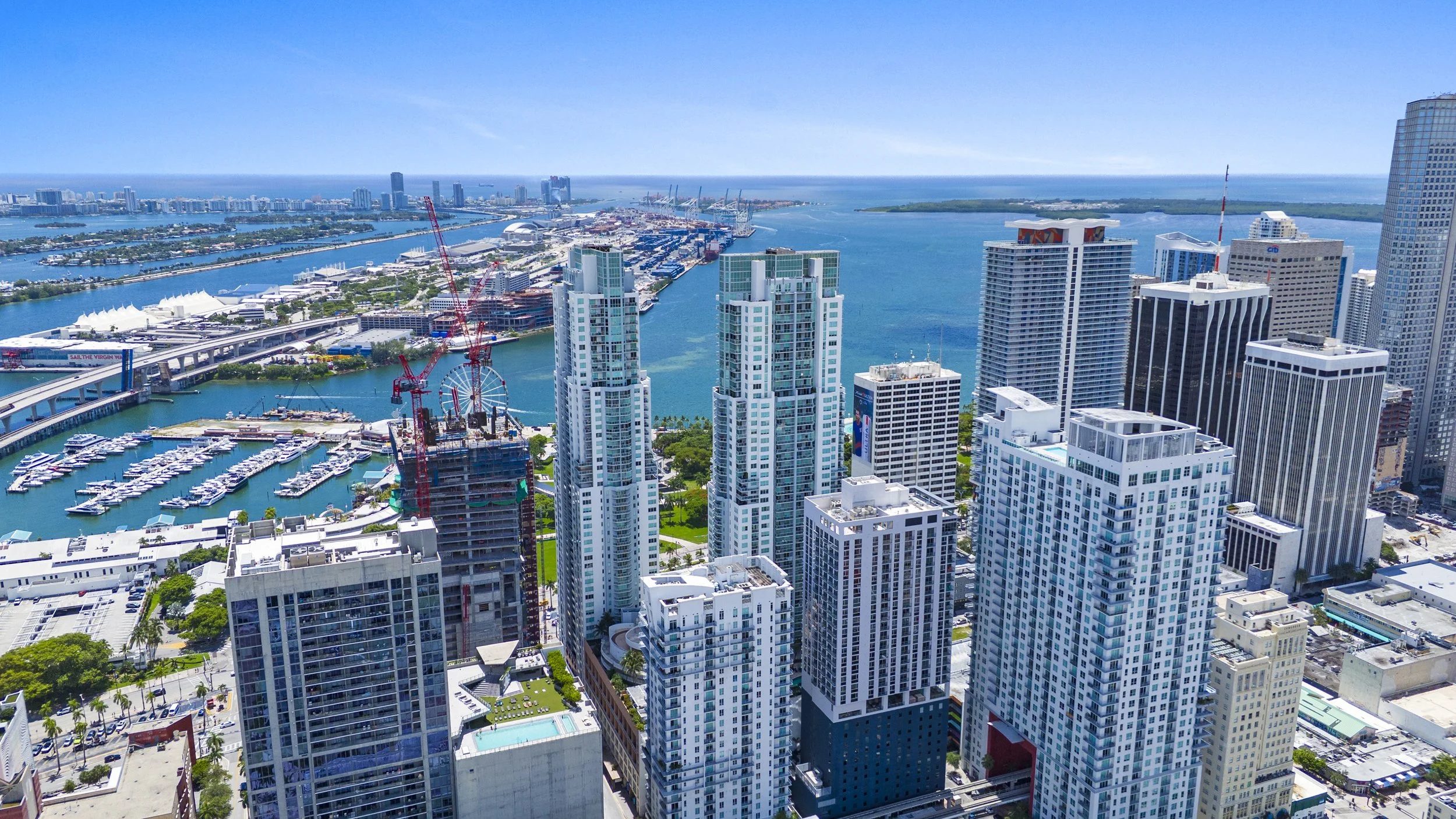 Aerial view of Miami skyline showing high-rise buildings, water channels, marina with boats, and distant cityscape under a bright blue sky.