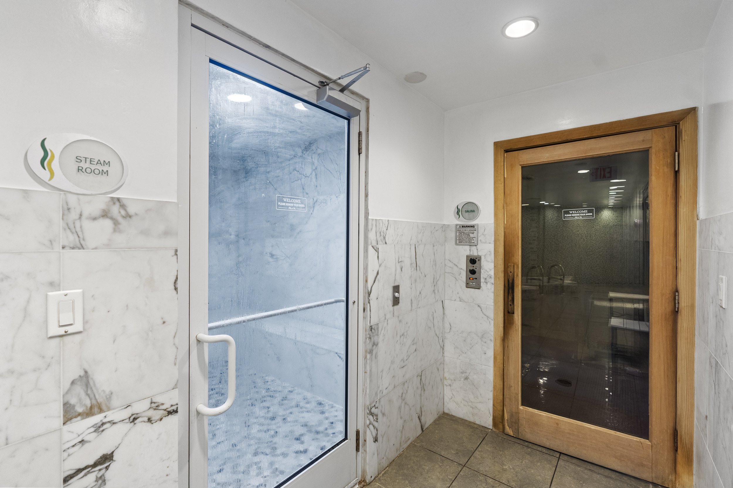 Indoor room with a glass door labeled "Steam Room" and a wooden door labeled "Sauna," both within a white-tiled wall space.