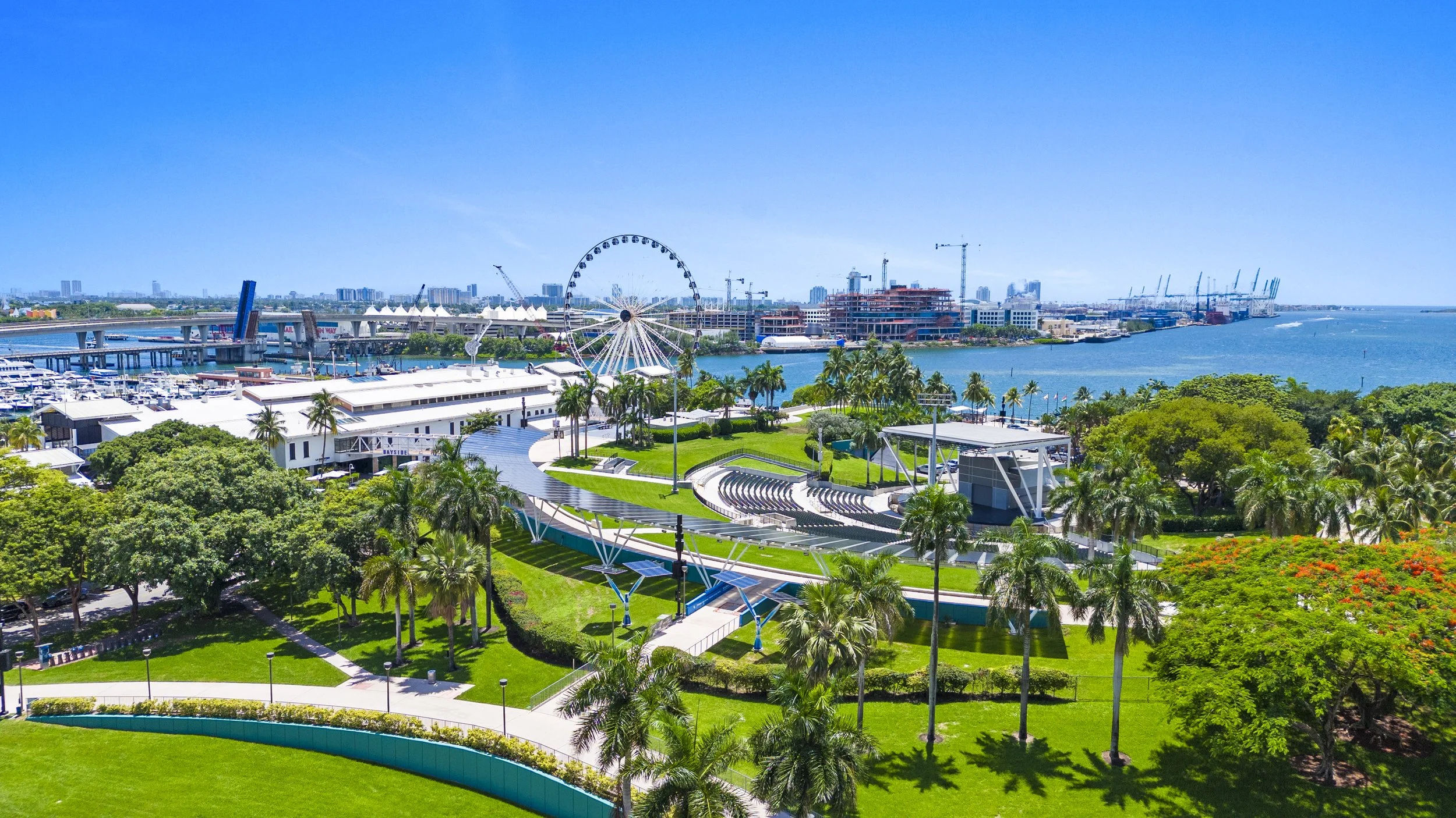A vibrant cityscape with a waterfront, amusement park rides including a Ferris wheel, lush green parks, palm trees, and a clear blue sky.