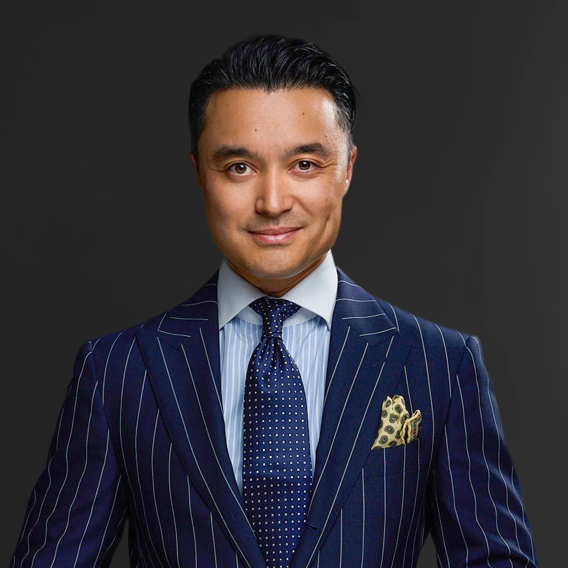 A man with dark hair and light skin, dressed in a dark blue pinstripe suit, light blue shirt, and navy tie with white polka dots, standing against a dark background.