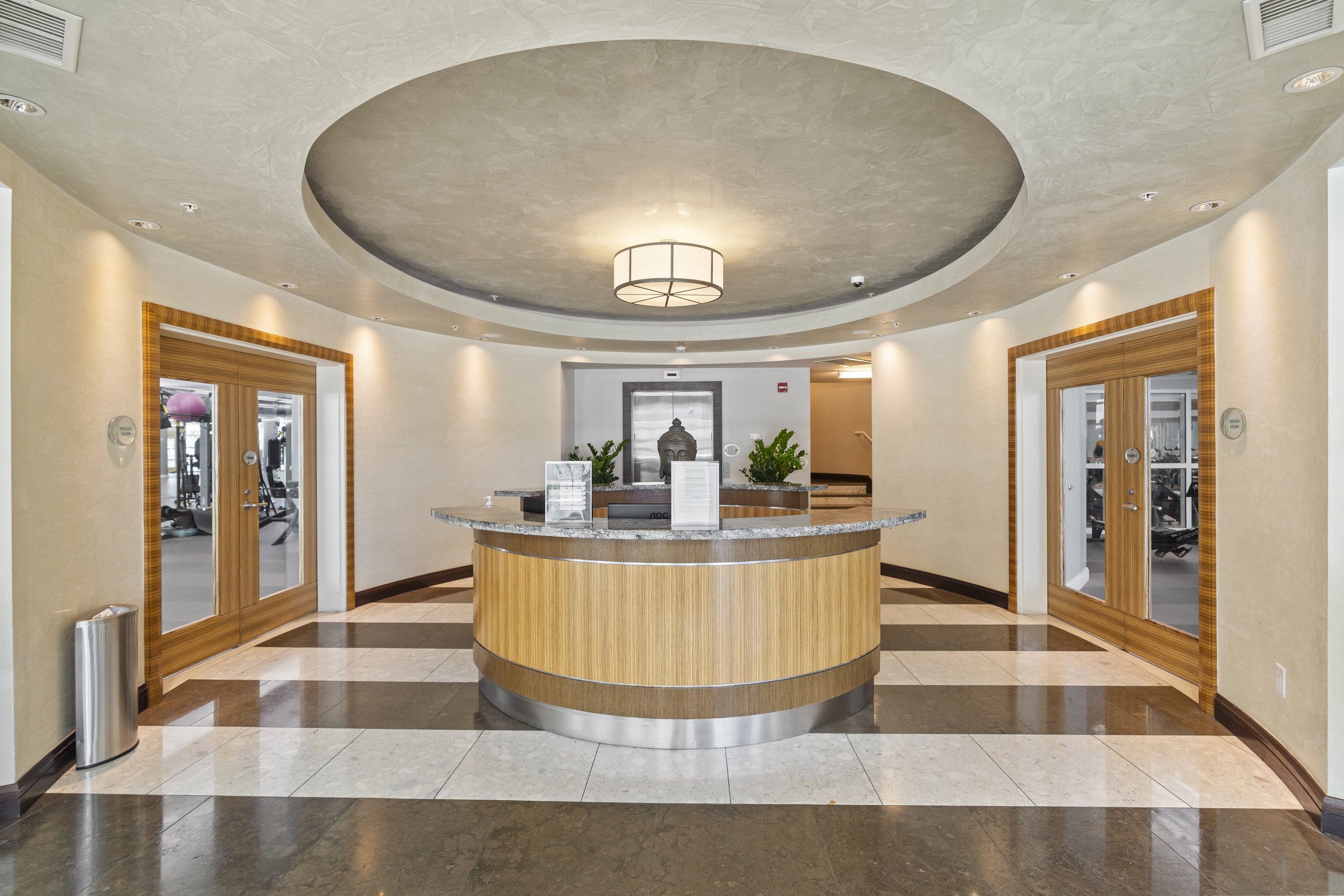 Hotel or office building reception desk with a circular wooden front, a Buddha statue, and green plants behind it. The ceiling is textured with a circular inset area and a modern chandelier. Two glass doors with wooden frames are on either side of th