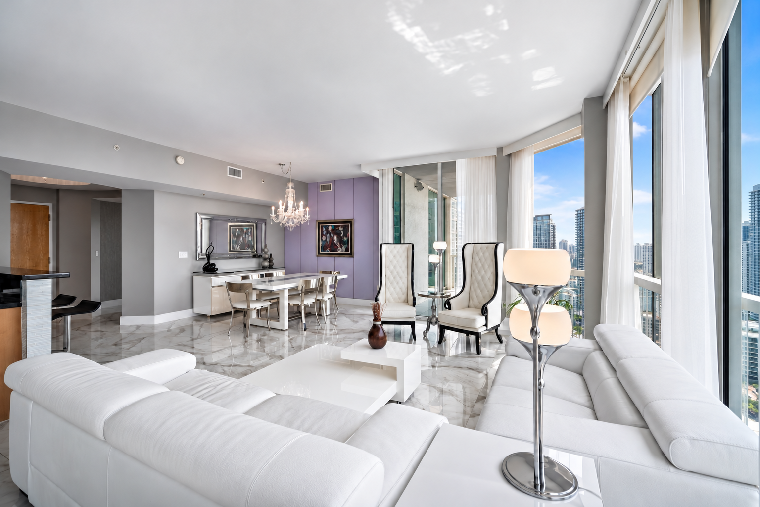 Luxury high-rise apartment living room with white sectional sofa, modern floor lamp, high-backed armchairs near large windows with city view, dining area with chandelier, marble floor, and contemporary artwork.