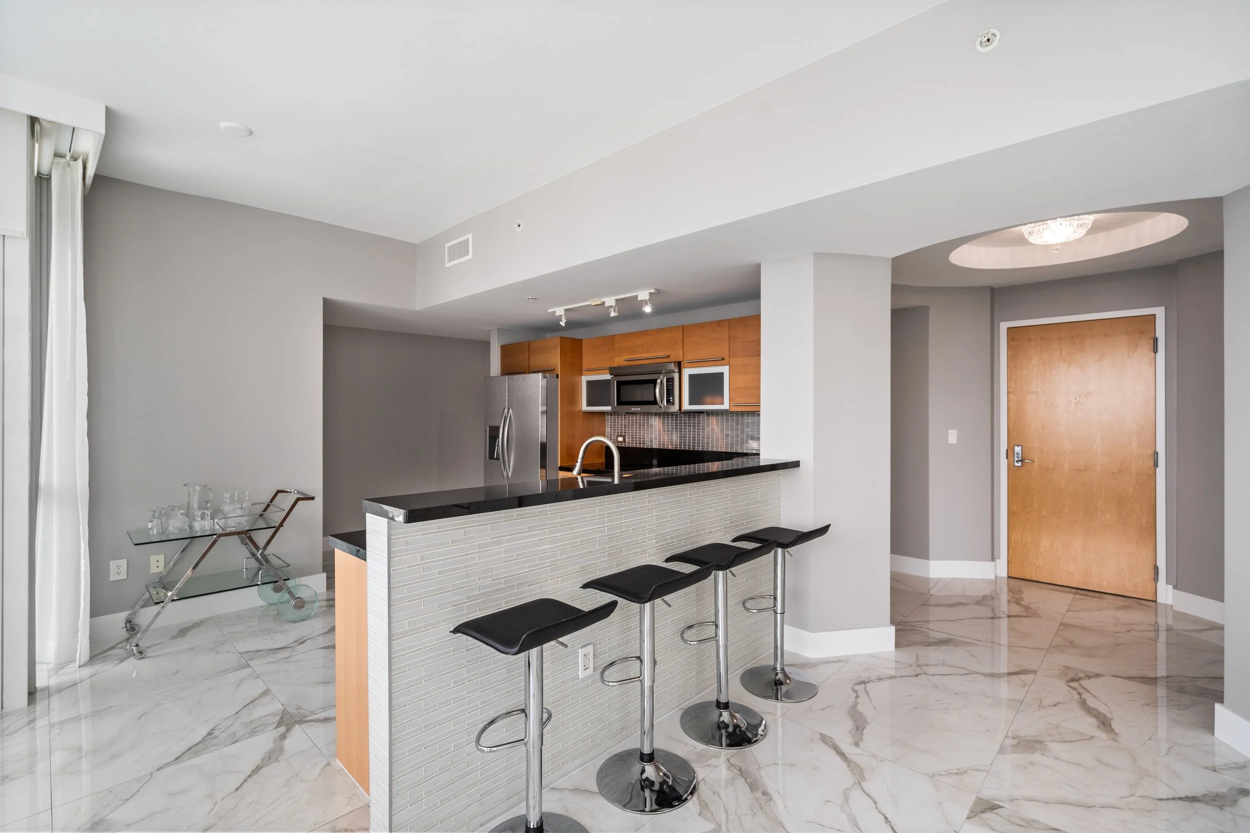 Modern kitchen with marble flooring, gray walls, wooden cabinets, stainless steel appliances, a breakfast bar with four black stools, and a chandelier visible through an archway.