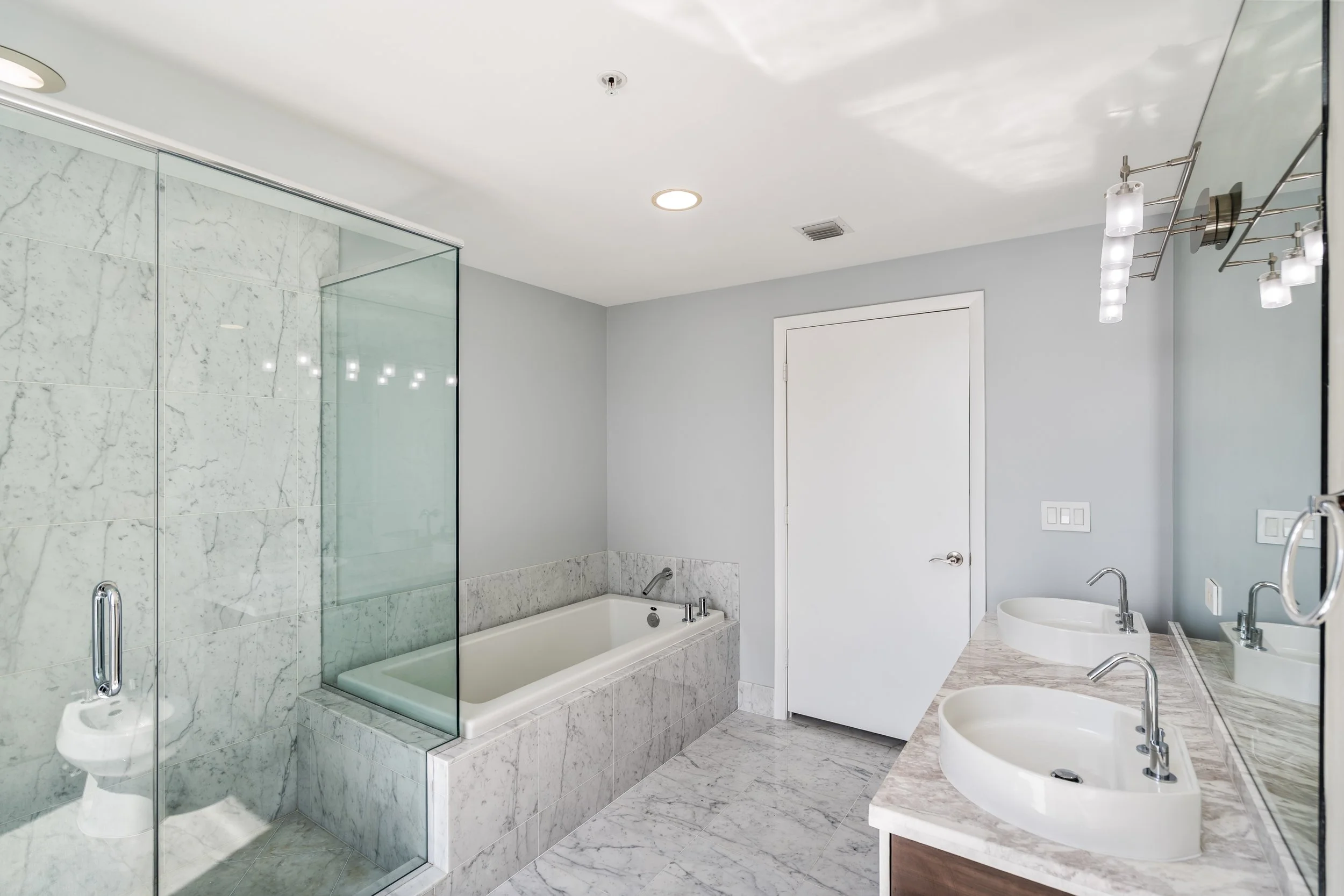 Modern bathroom with a glass-enclosed shower, marble tub, white double sinks, and light gray walls.