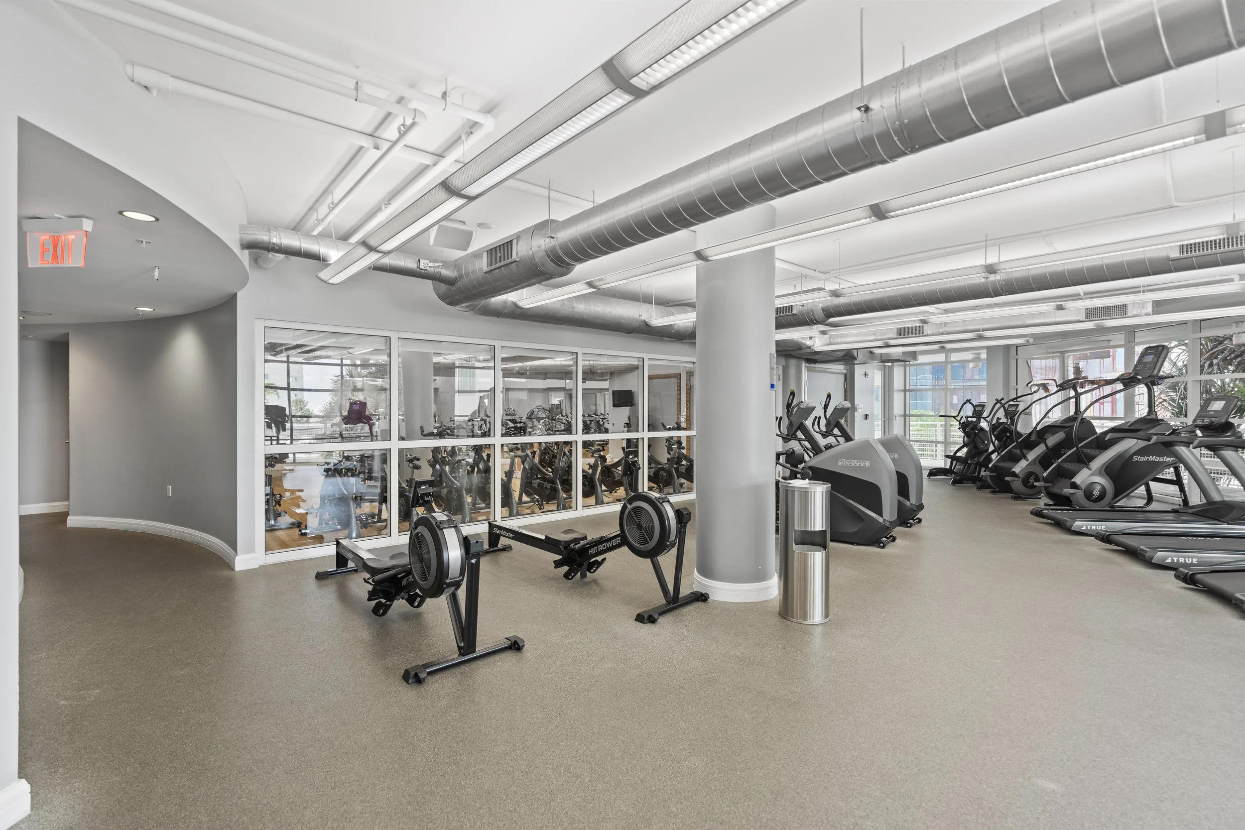Indoor gym with cardio equipment, including treadmills, elliptical machines, and a rowing machine, separated by glass walls with a view of another workout area.