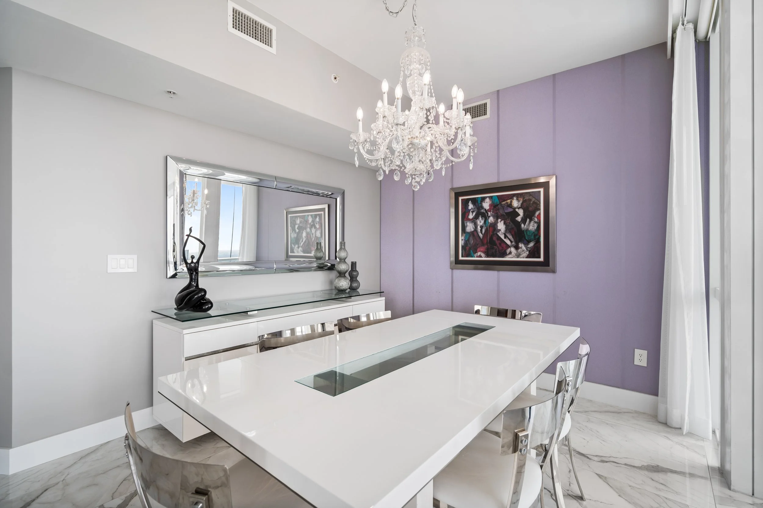 Modern dining room with white table, silver chairs, purple accent wall, chandelier, colorful abstract artwork, and decorative vases.
