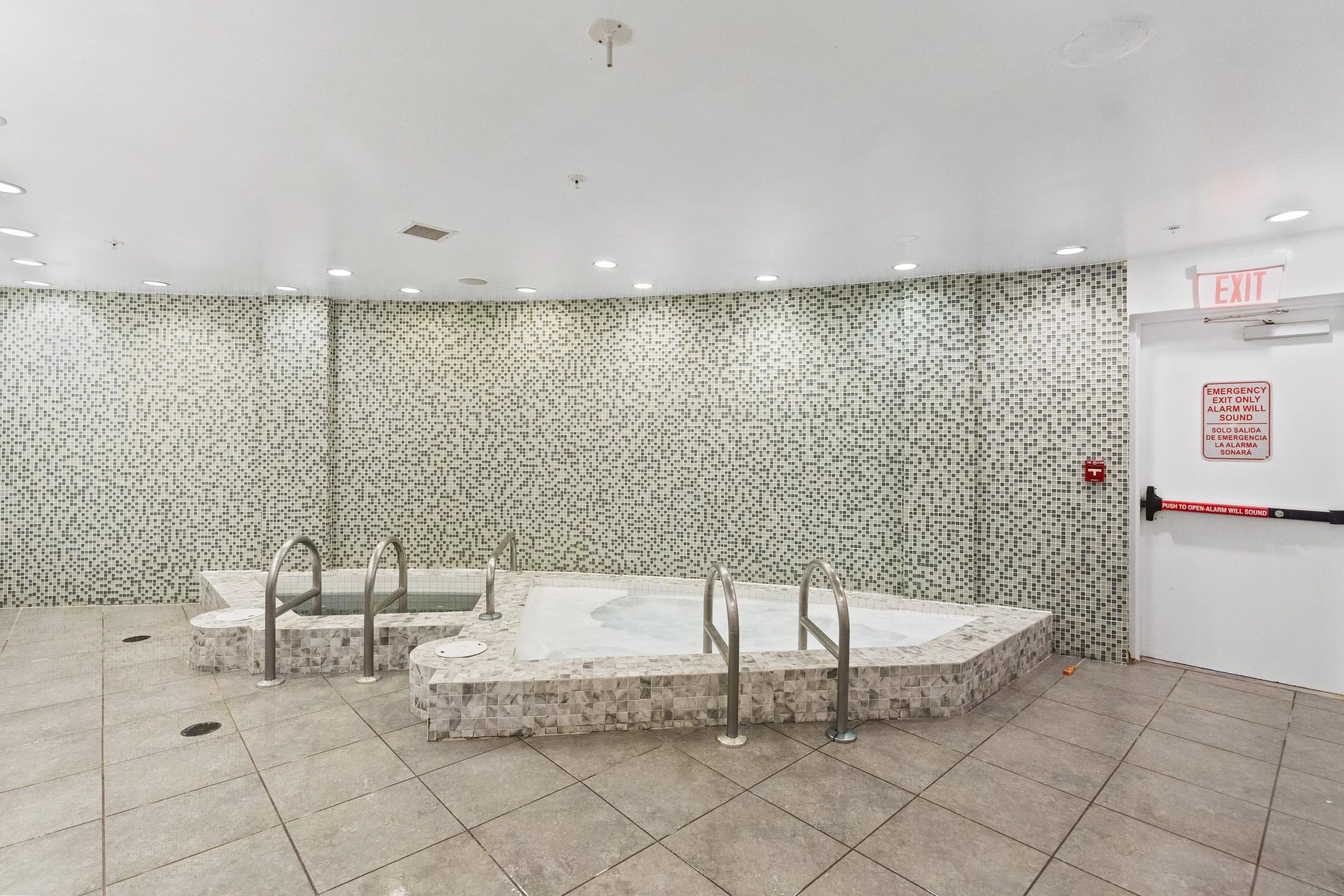 Indoor hot tub area with tiled walls, stainless steel handrails, and a white ceiling with recessed lighting. There is a red emergency exit sign and a fire alarm on the wall, and the hot tub is surrounded by a tiled platform.