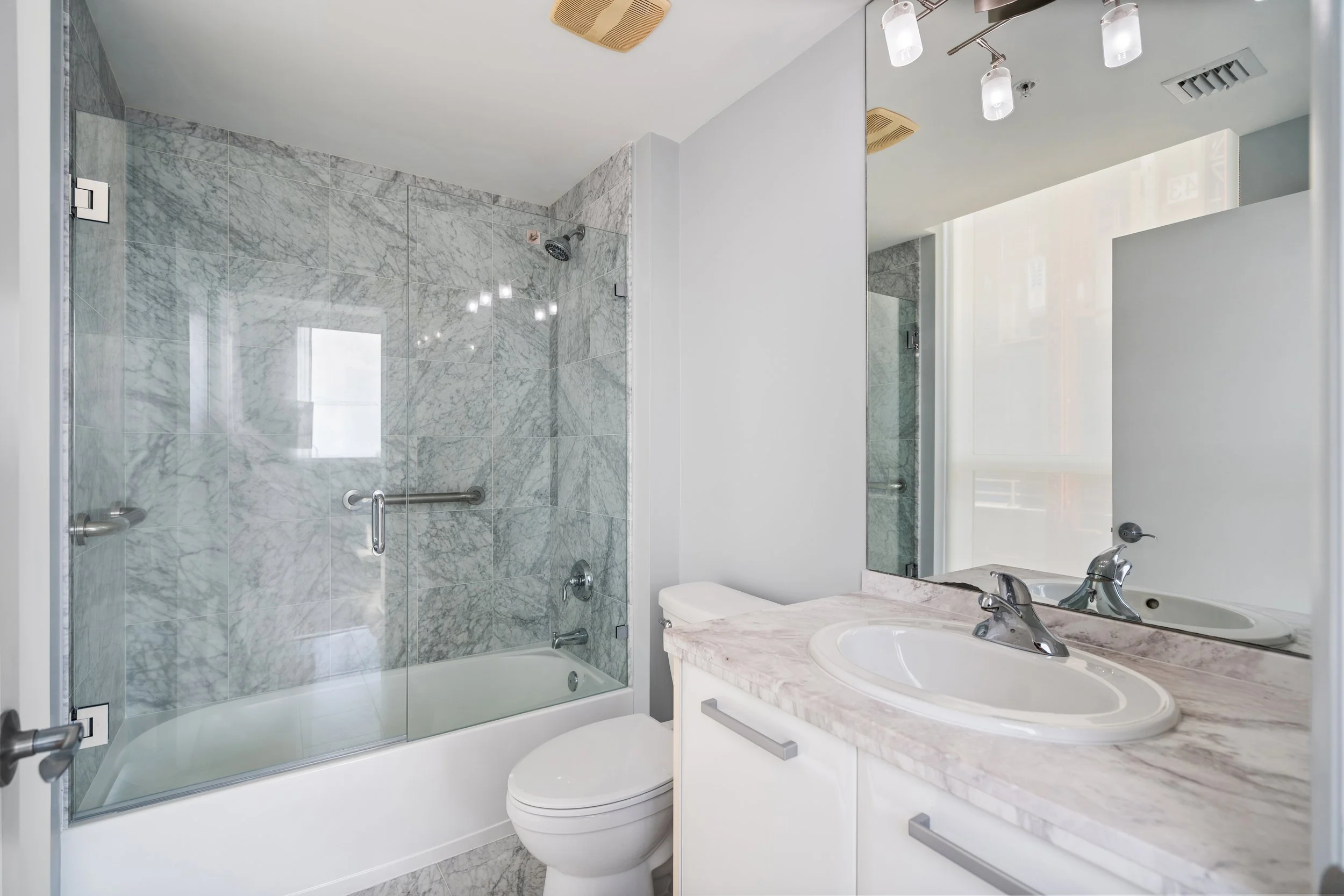Modern bathroom with marble shower enclosure, white toilet, and marble countertop with sink, large mirror, and ceiling light fixtures.