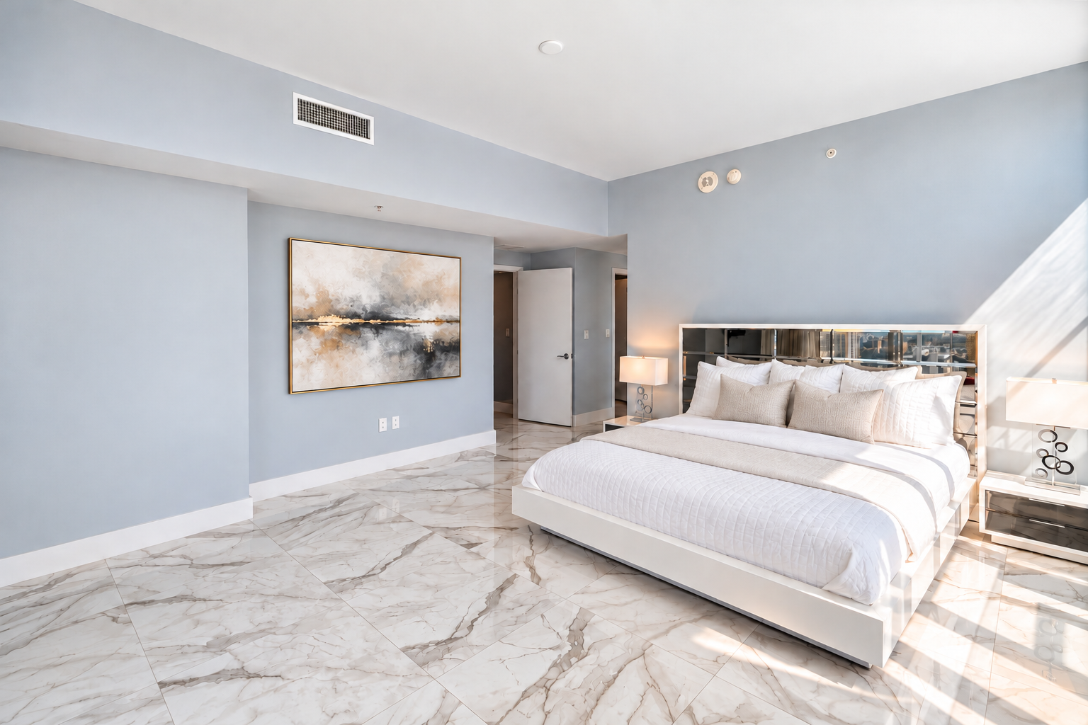 Modern bedroom with white bed, beige pillows, marble floor, blue walls, artwork, and bedside lamps.