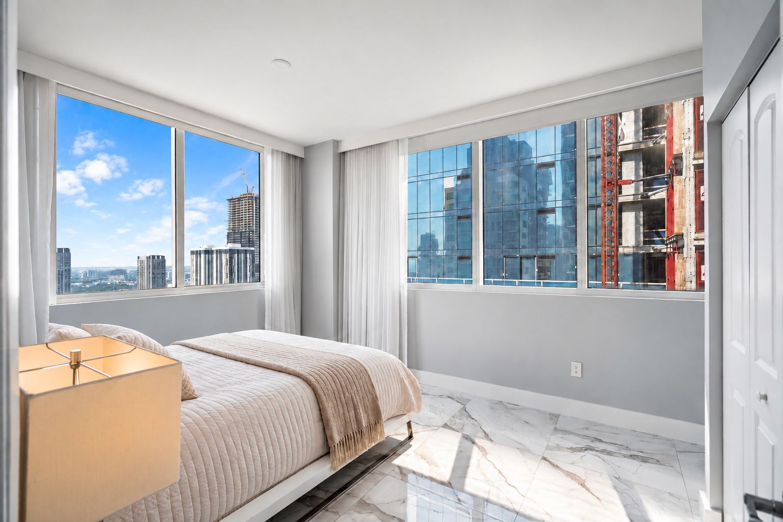 Bright bedroom with large windows, city skyline view, bed with light pink bedding, marble floor, and white walls.