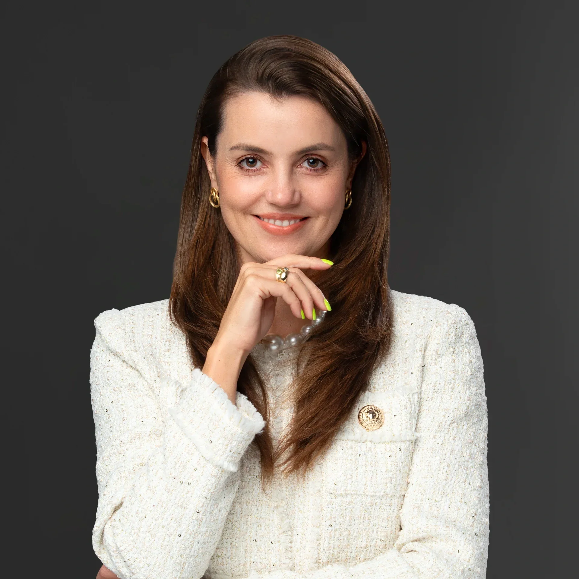 A woman with long brown hair, wearing gold earrings, a pearl necklace, and a cream-colored blazer with a badge, smiling and resting her chin on her hand with painted neon yellow nails, against a dark gray background.