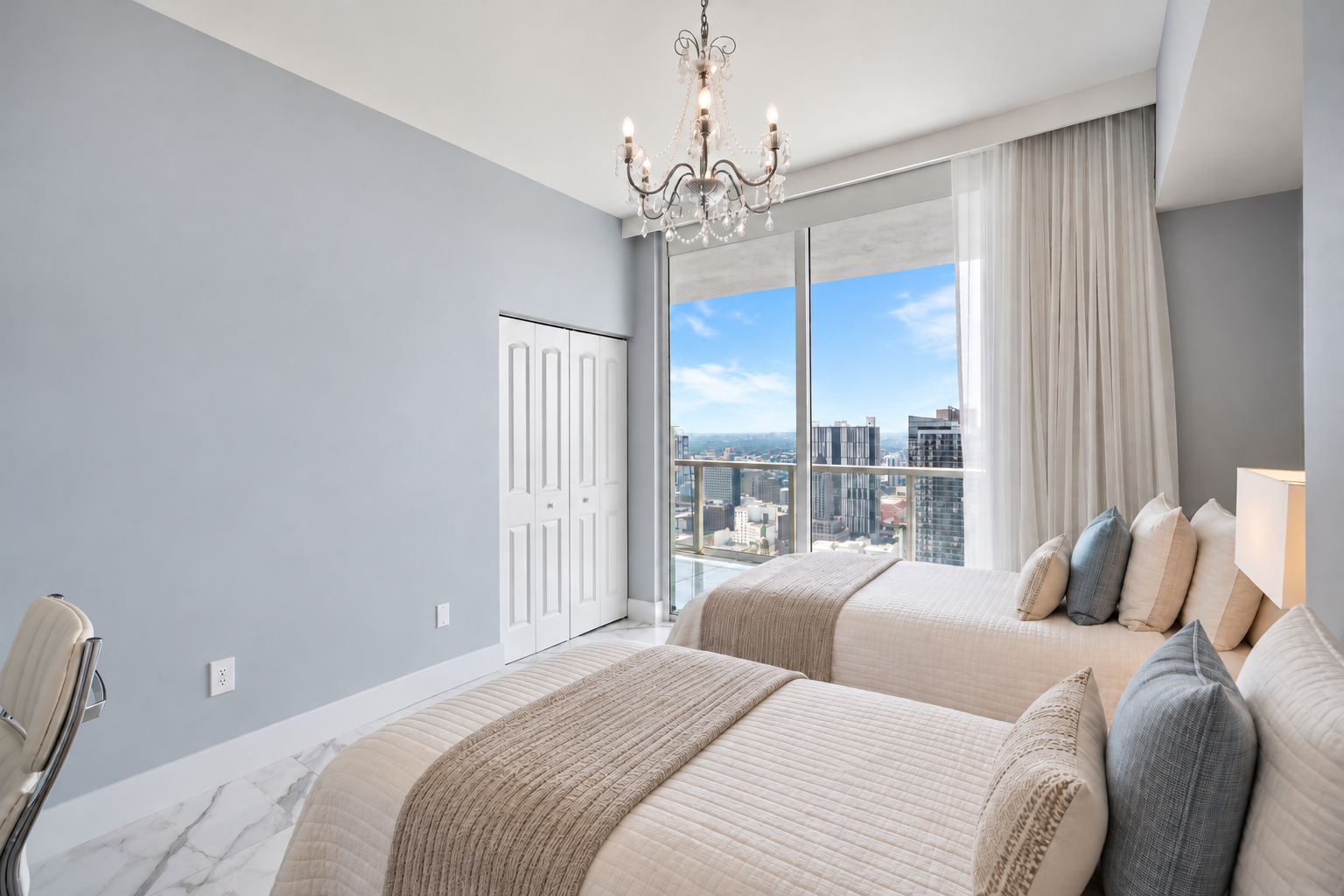 A modern bedroom with two twin beds, a large window showing city skyscrapers and a blue sky, a chandelier hanging from the ceiling, and white marble flooring.
