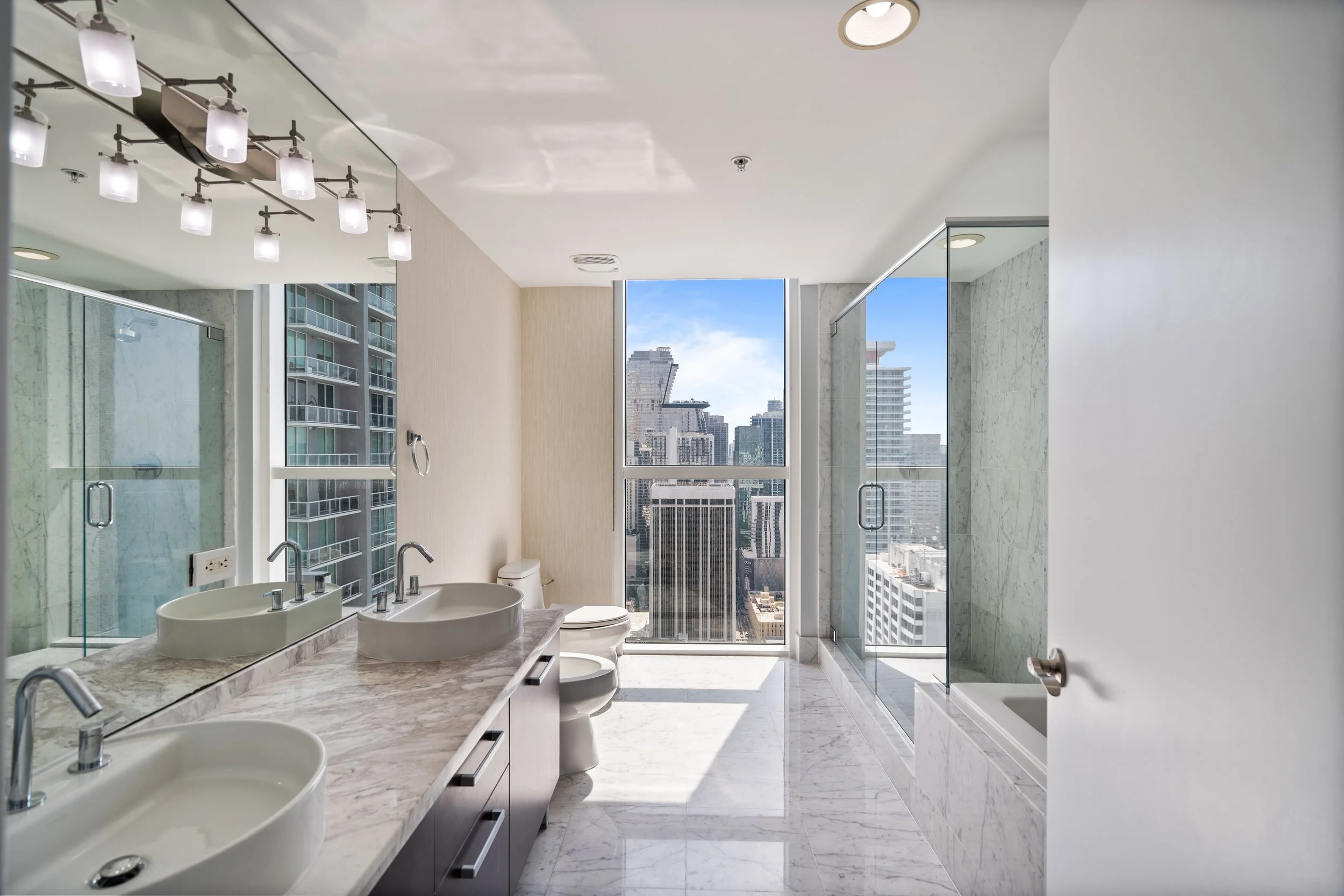 Modern bathroom with marble floors and walls, double sinks with a marble countertop, large mirror, city view through big windows, glass shower enclosure, and contemporary lighting fixtures.
