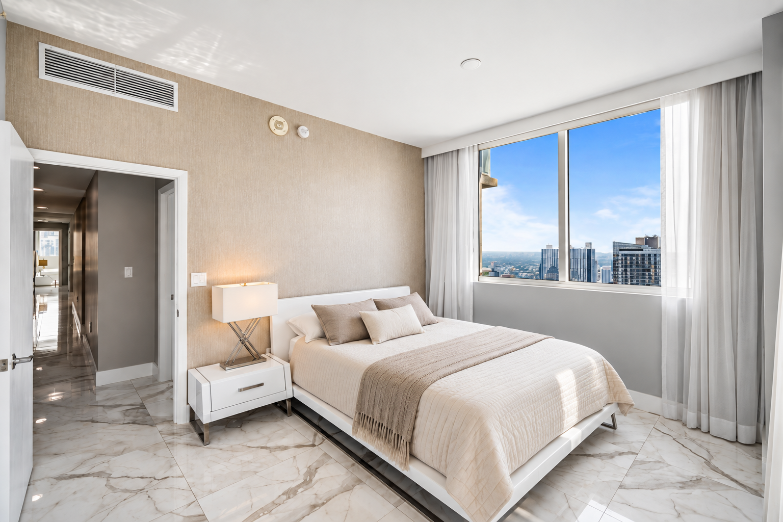 Modern bedroom with large window view of city skyline, beige walls, white bed with neutral pillows, marble floor, and white nightstand with lamp.