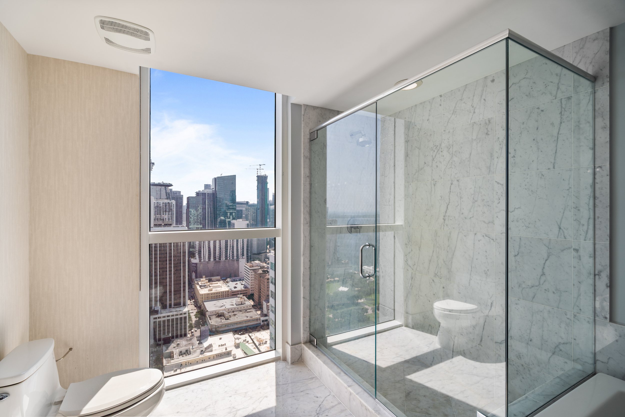 Modern bathroom with a glass shower enclosure, white toilet, marble floor, large window with cityscape view, beige and gray walls.