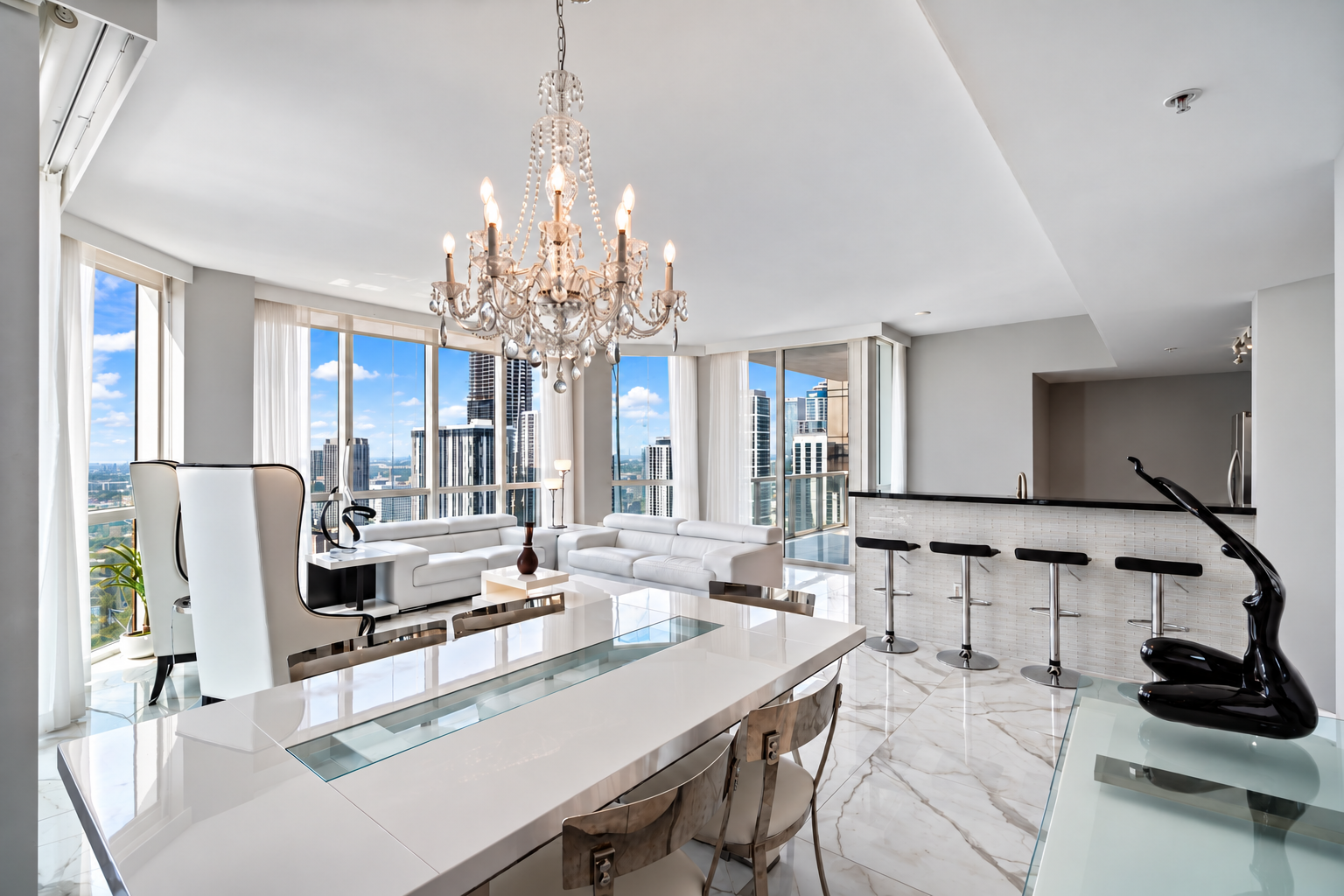 Modern high-rise apartment living room with large windows, white sofas, a dining table, chandelier, bar area with stools, and city skyline view.
