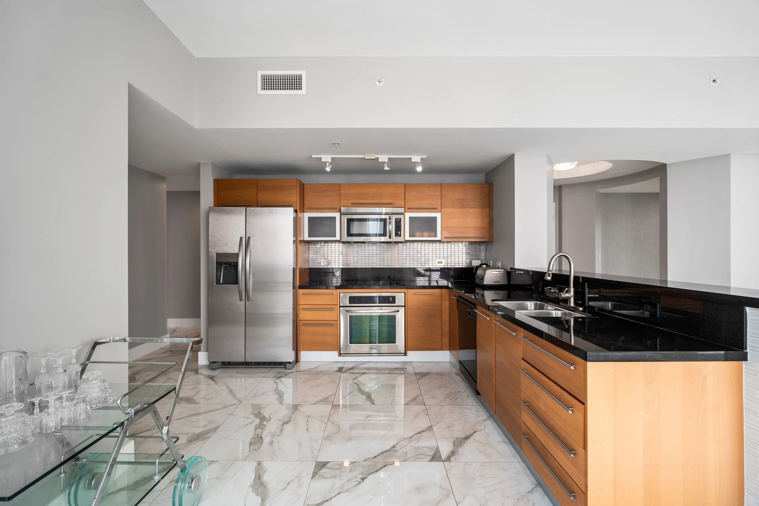 Modern kitchen with stainless steel refrigerator, built-in microwave, oven, black granite countertops, wooden cabinets, marble floor, and a dining area with glass table and clear chairs.