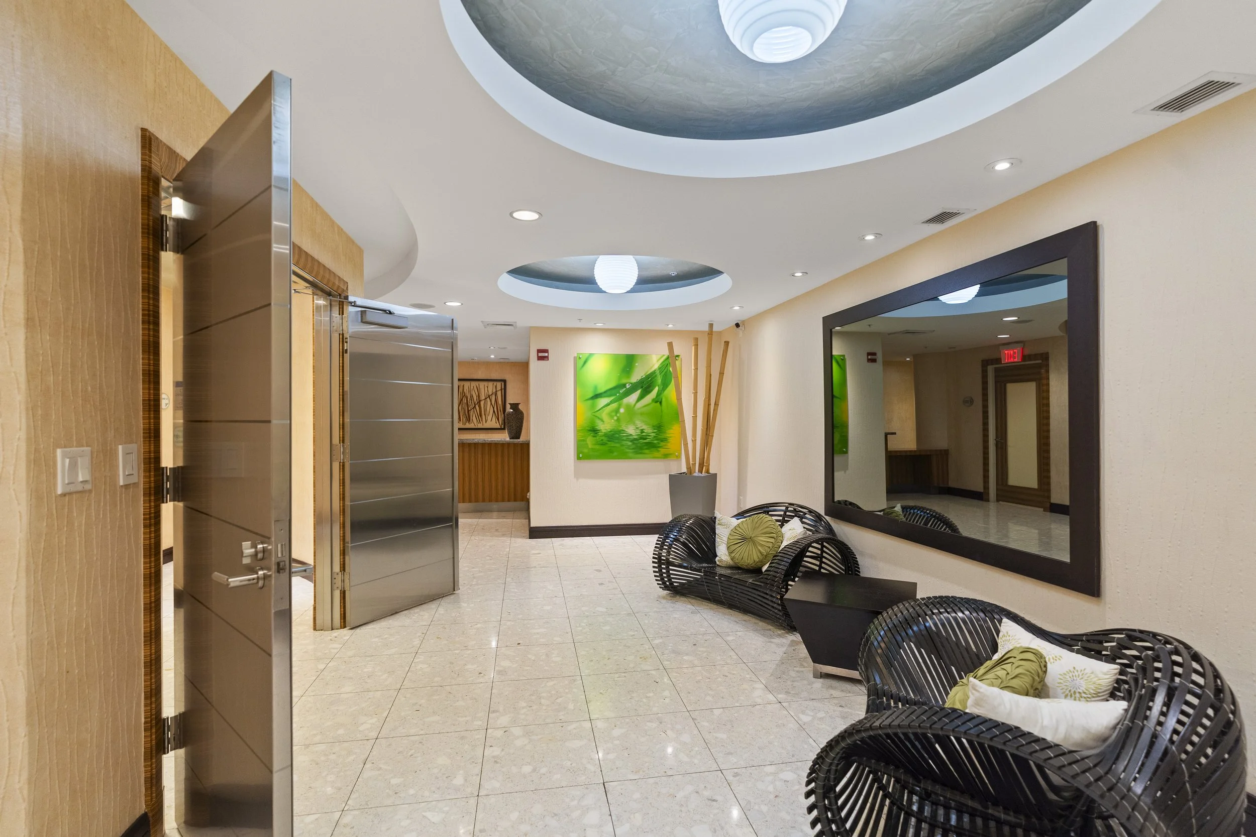 Lobby area with black chairs and pillows, a large mirror, a green floral painting on the wall, bamboo decor, and ceiling lights.