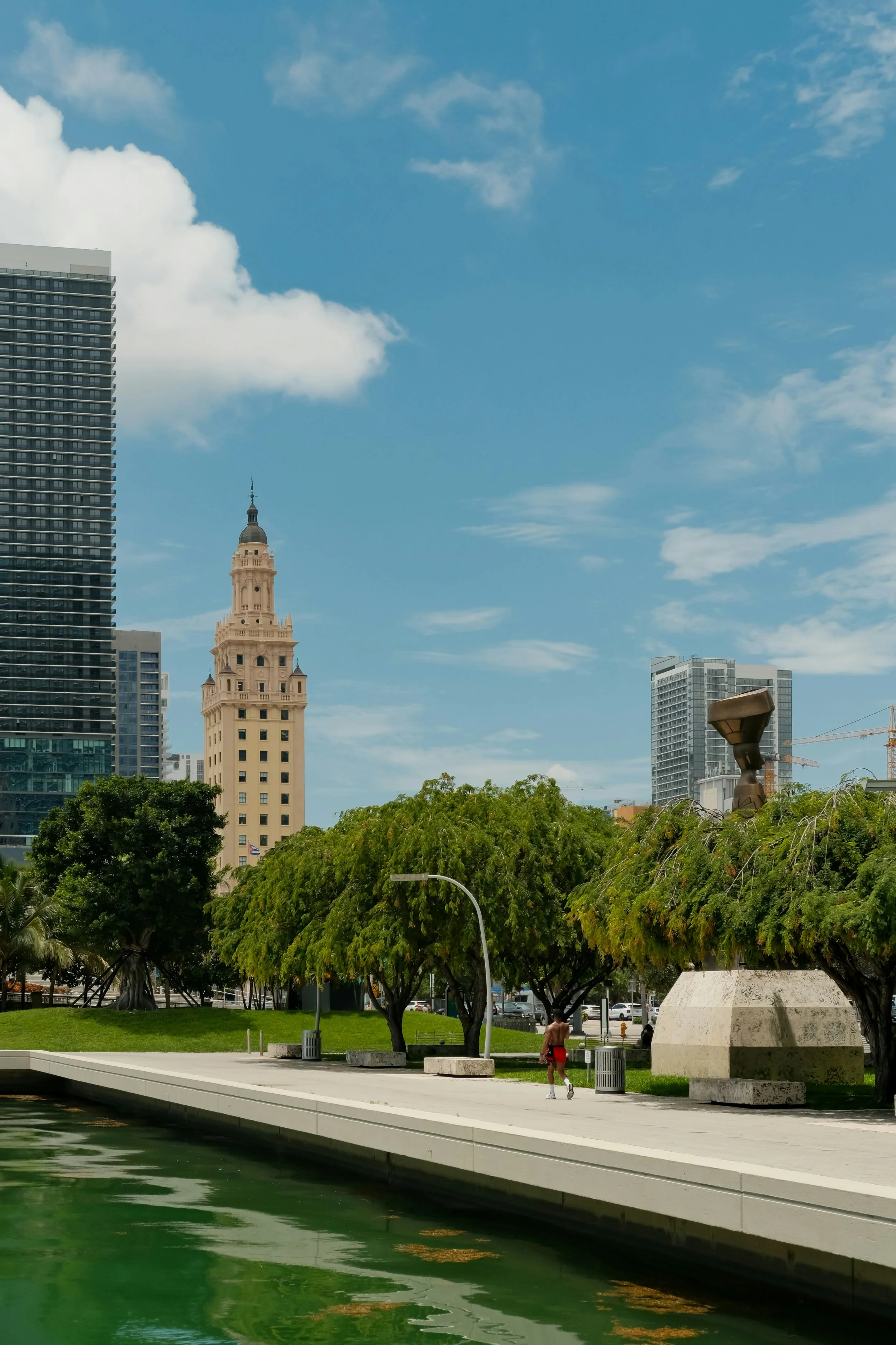 Cityscape with a river in the foreground, lined with a sidewalk and trees. Tall buildings, including a historic clock tower and modern skyscrapers, are seen against a partly cloudy sky.
