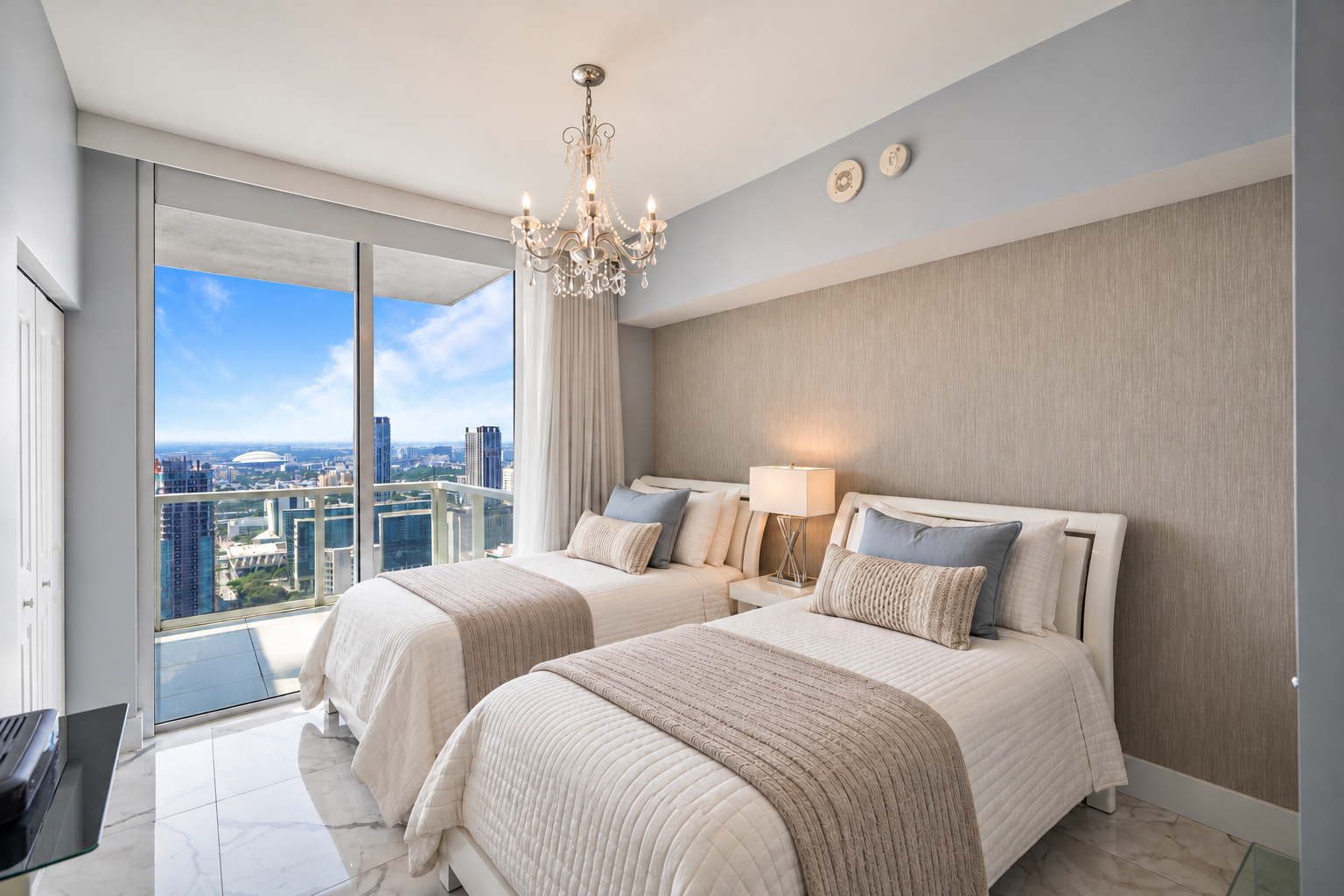 A modern bedroom with two white beds, beige and blue pillows, a bedside lamp, and a chandelier, with a large glass sliding door opening to a balcony with a city view of skyscrapers and a stadium under a blue sky.