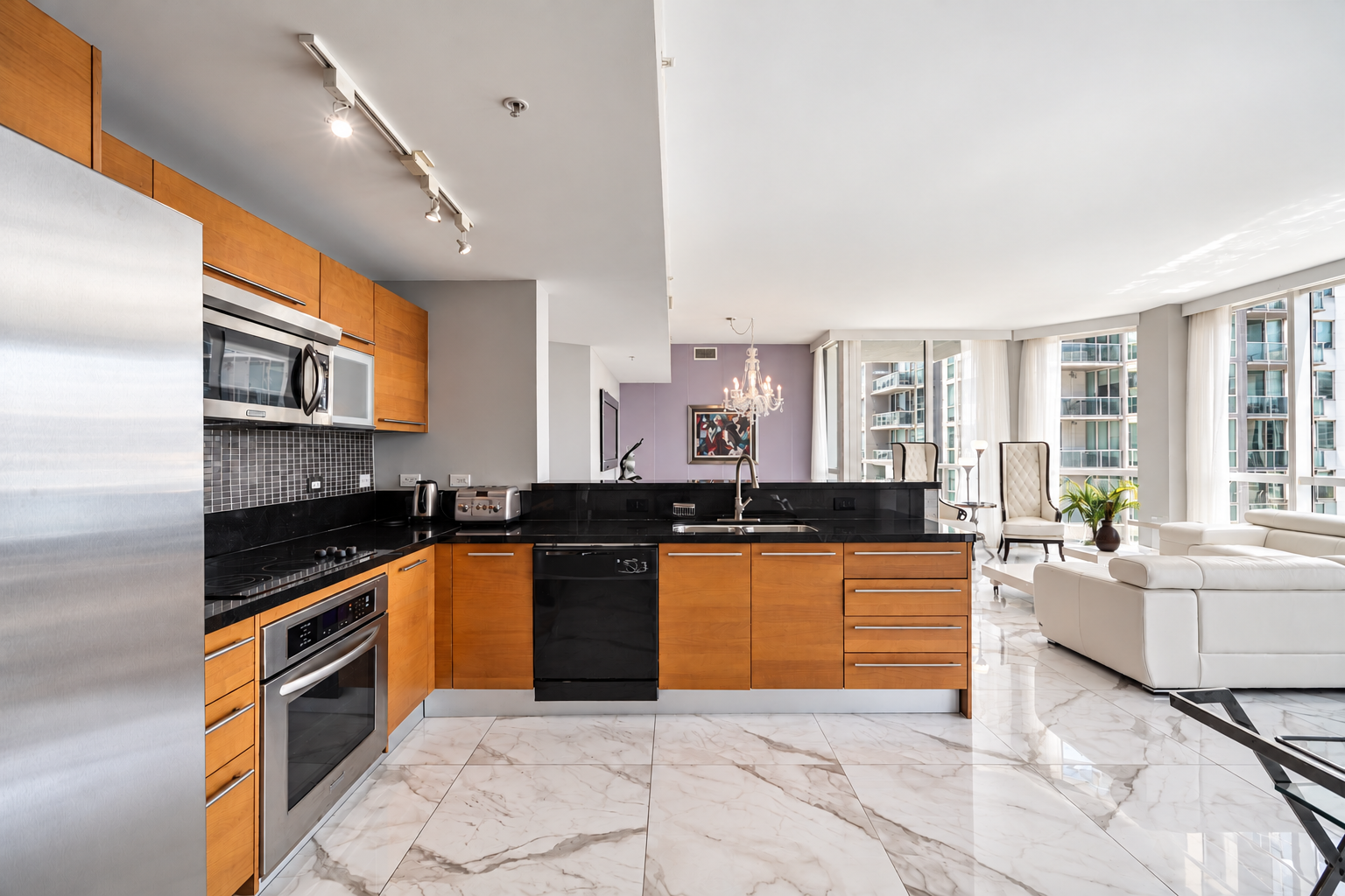 Modern open-concept kitchen with wooden cabinets, black countertops, stainless steel appliances, and a view of a bright living room with large windows and white furniture.