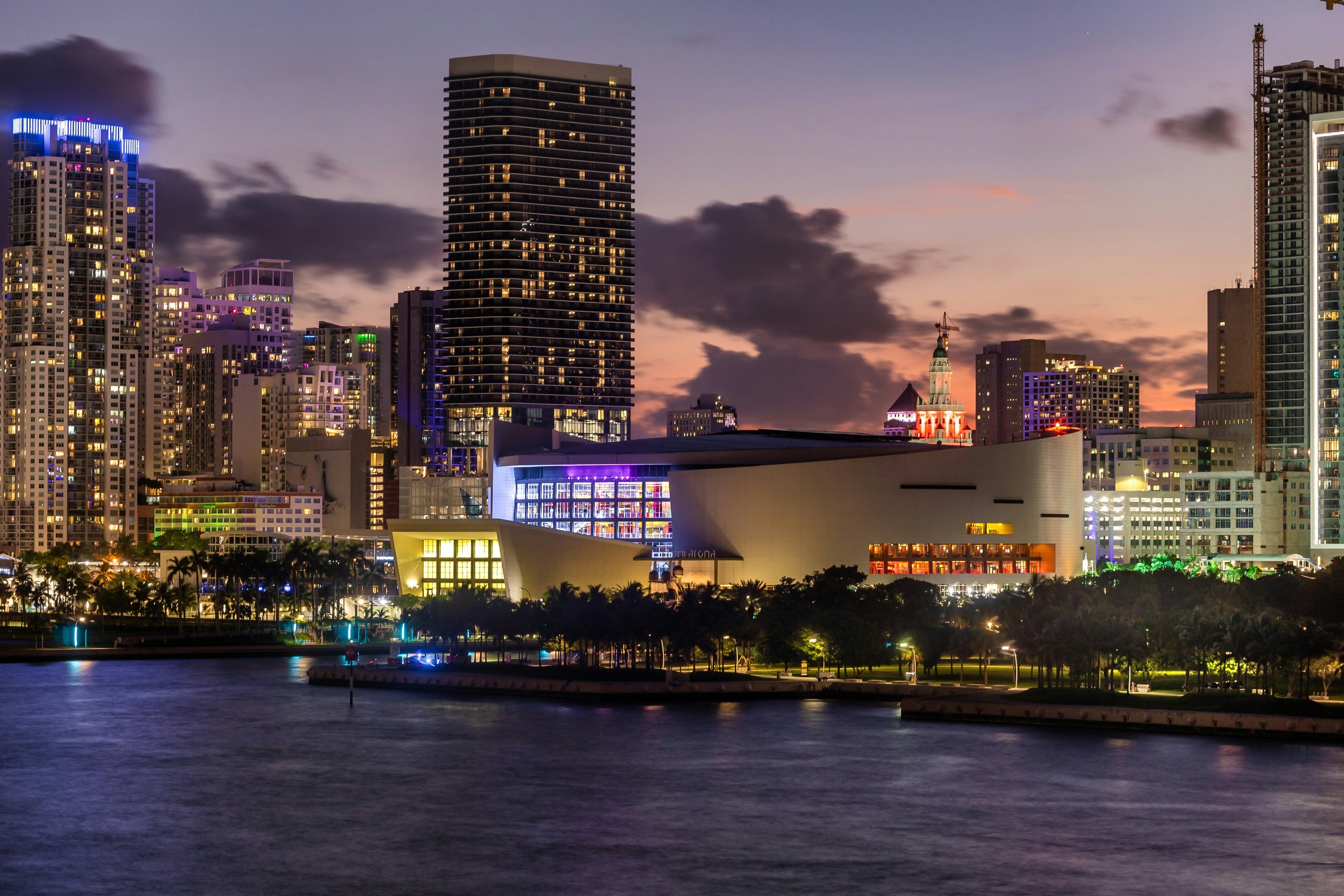 City skyline at dusk with tall buildings, illuminated windows, and a body of water in the foreground.
