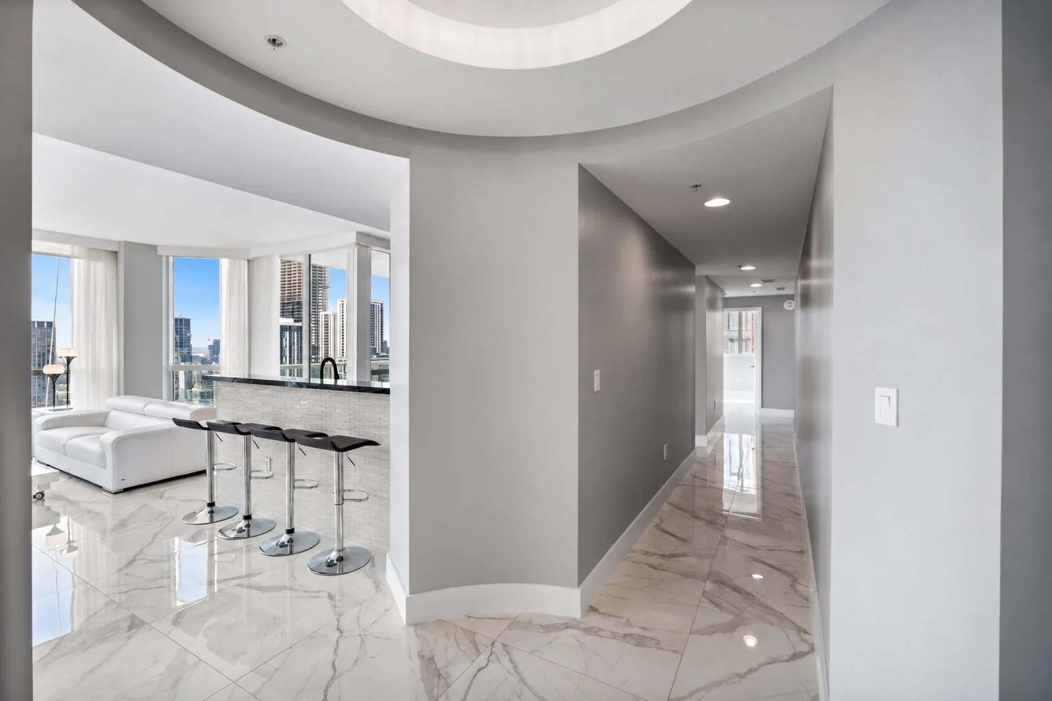 Modern apartment interior with marble flooring, a hallway leading to a sunset view, and a living area with a white sofa and bar stools near large windows showing a cityscape.