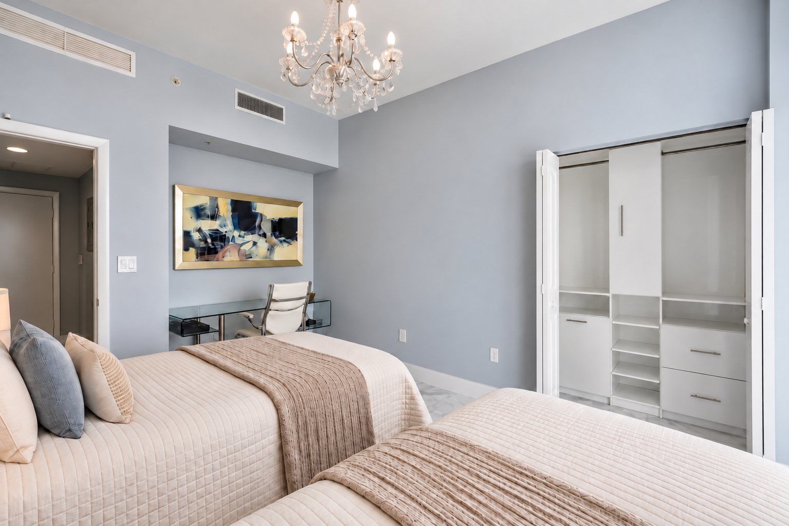 A bedroom with two beige beds, a glass desk with a white chair, an abstract painting on the wall, a chandelier, and an empty white closet with sliding doors.