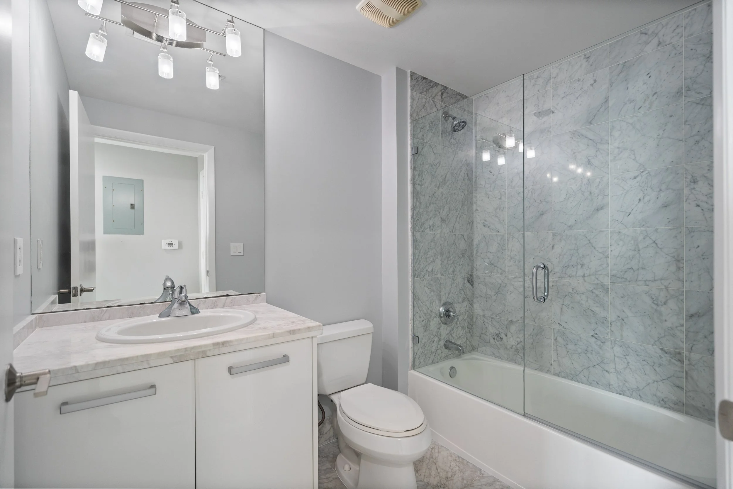 Modern bathroom with white vanity, marble countertop, and a glass shower enclosure with marble tiles. Overhead lighting, mirror, and a toilet are visible.