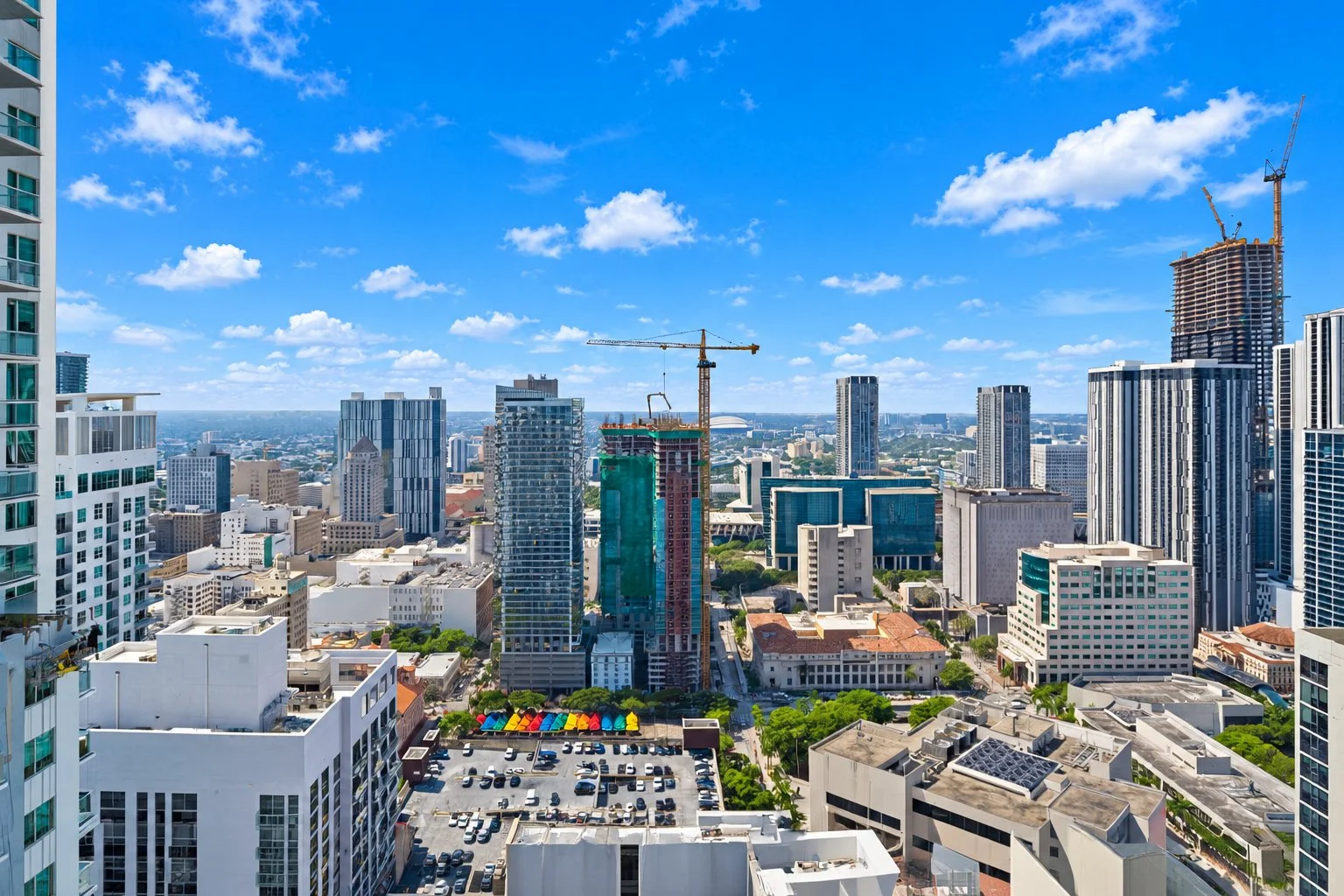 City skyline with tall buildings, construction cranes, and a partly cloudy blue sky.