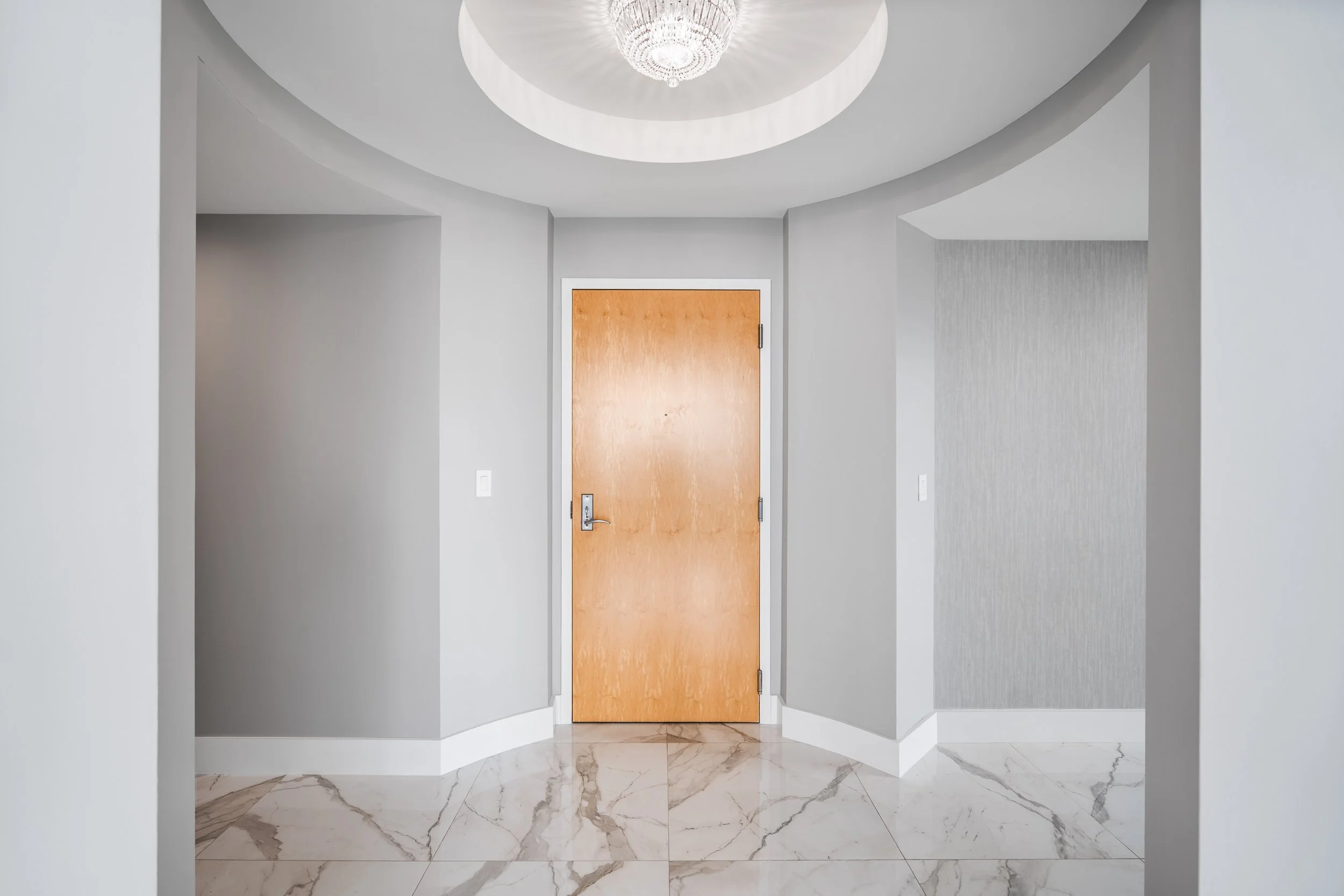 Interior hallway with white walls, a wooden door at the end, marble flooring with gray and white veining, and a circular recessed ceiling with a chandelier.