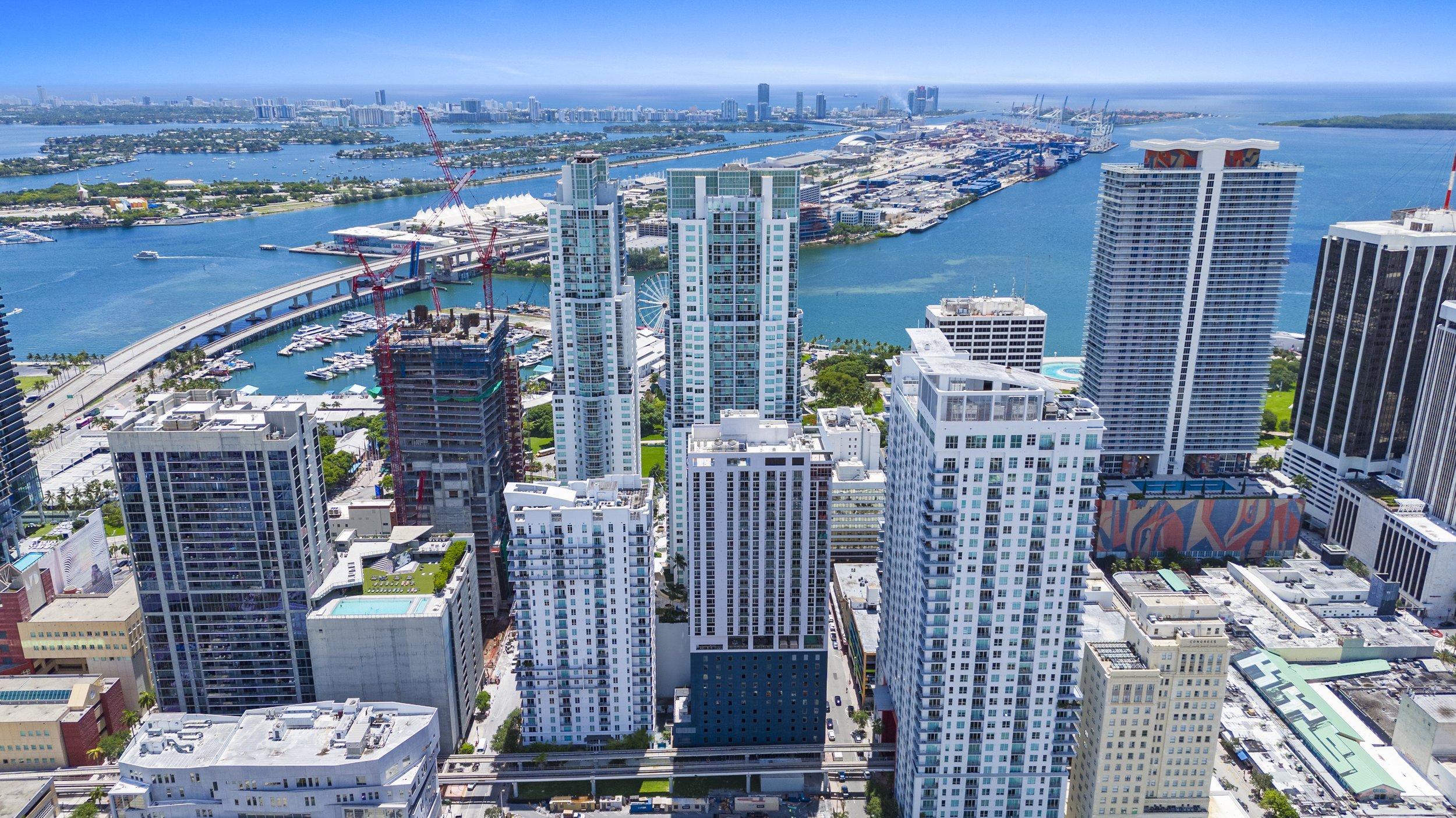 Aerial view of a city skyline with tall modern buildings, waterways, and a harbor with cranes and ships, possibly Miami, Florida.