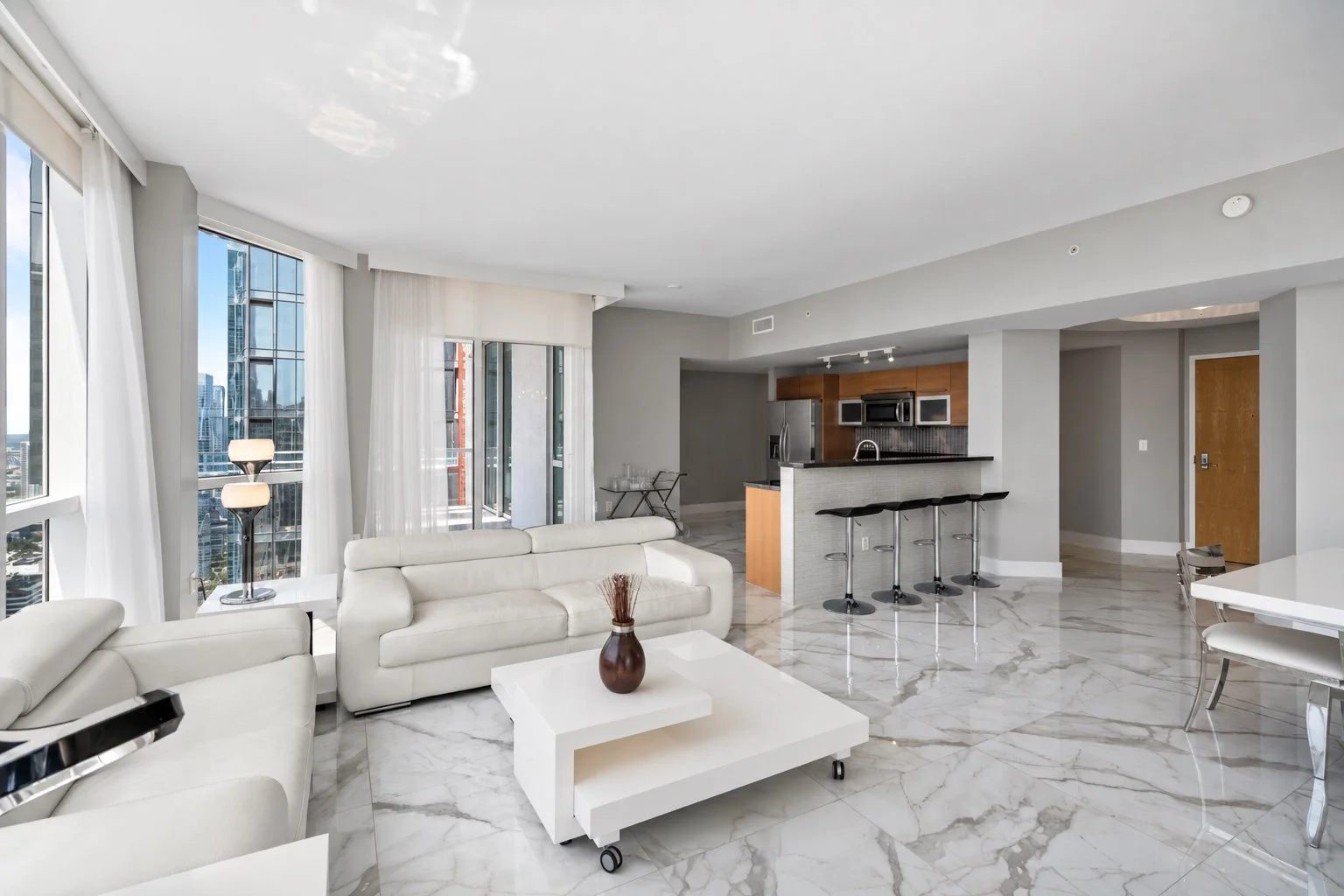 Modern apartment living room with white leather sofas, marble flooring, large windows with city views, a small bar area with barstools, and a kitchen with wooden cabinets and stainless steel appliances.