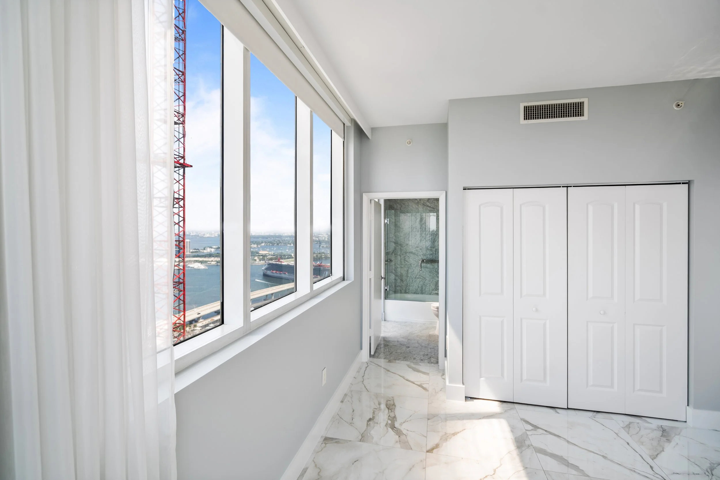 Interior of a modern high-rise apartment with large windows showing city and water views, white marble floors, and white double closet doors.