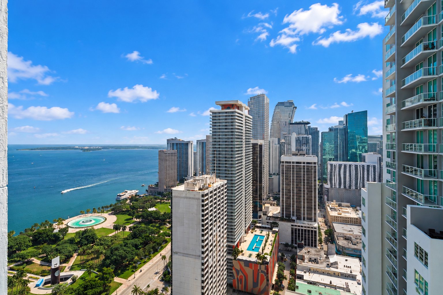 A city skyline featuring tall modern skyscrapers, a waterfront with boats, and a green park with a water fountain, all under a bright blue sky with scattered clouds.