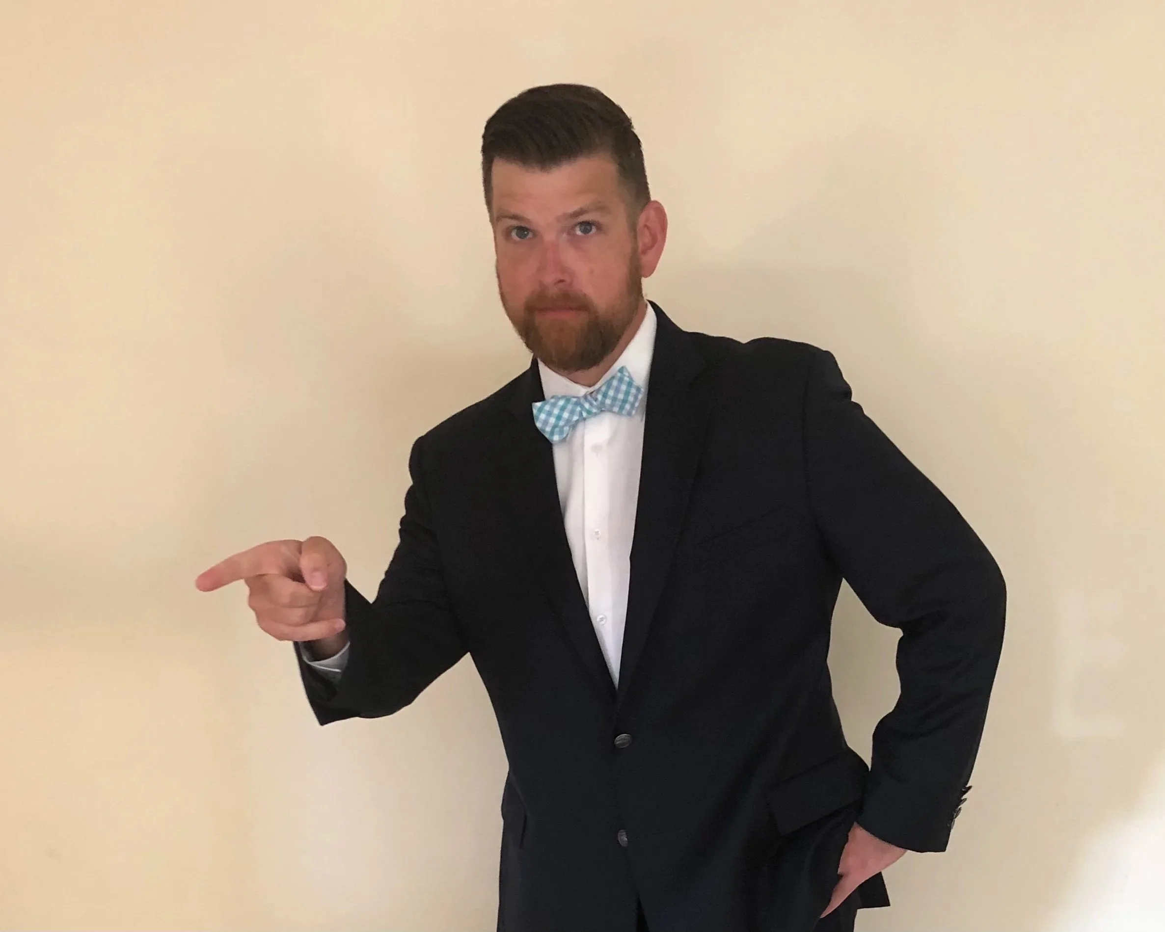 A man in a tuxedo with a light blue checkered bow tie, pointing with his right hand, standing in front of a plain off-white wall.