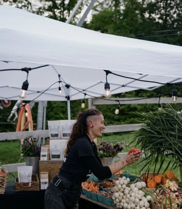 Femme vendant des légumes frais à un stand en plein air sous une tente blanche avec des guirlandes lumineuses
