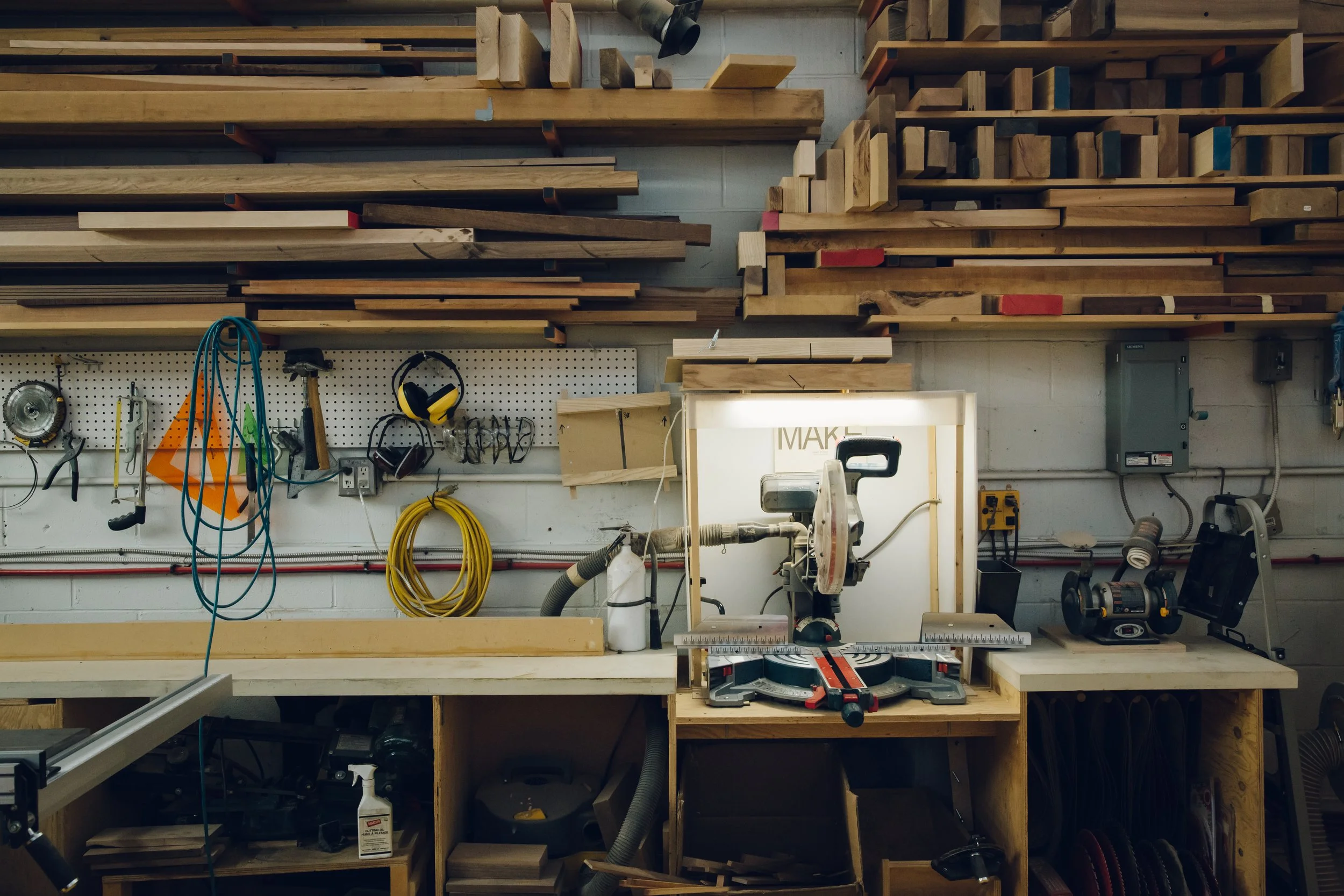 A woodworking workshop with wooden shelves holding various sizes of lumber, tools, and equipment. The workspace includes power tools, a workbench, and organized storage for woodworking supplies.