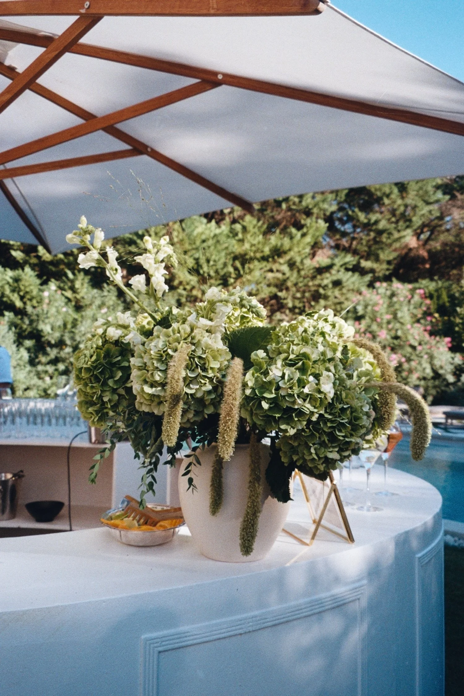 Un grand vase blanc avec des fleurs vertes, blanches et des longues clochettes suspendues, placé sur une table extérieure blanche sous un parasol blanc. Décoration de mariage
