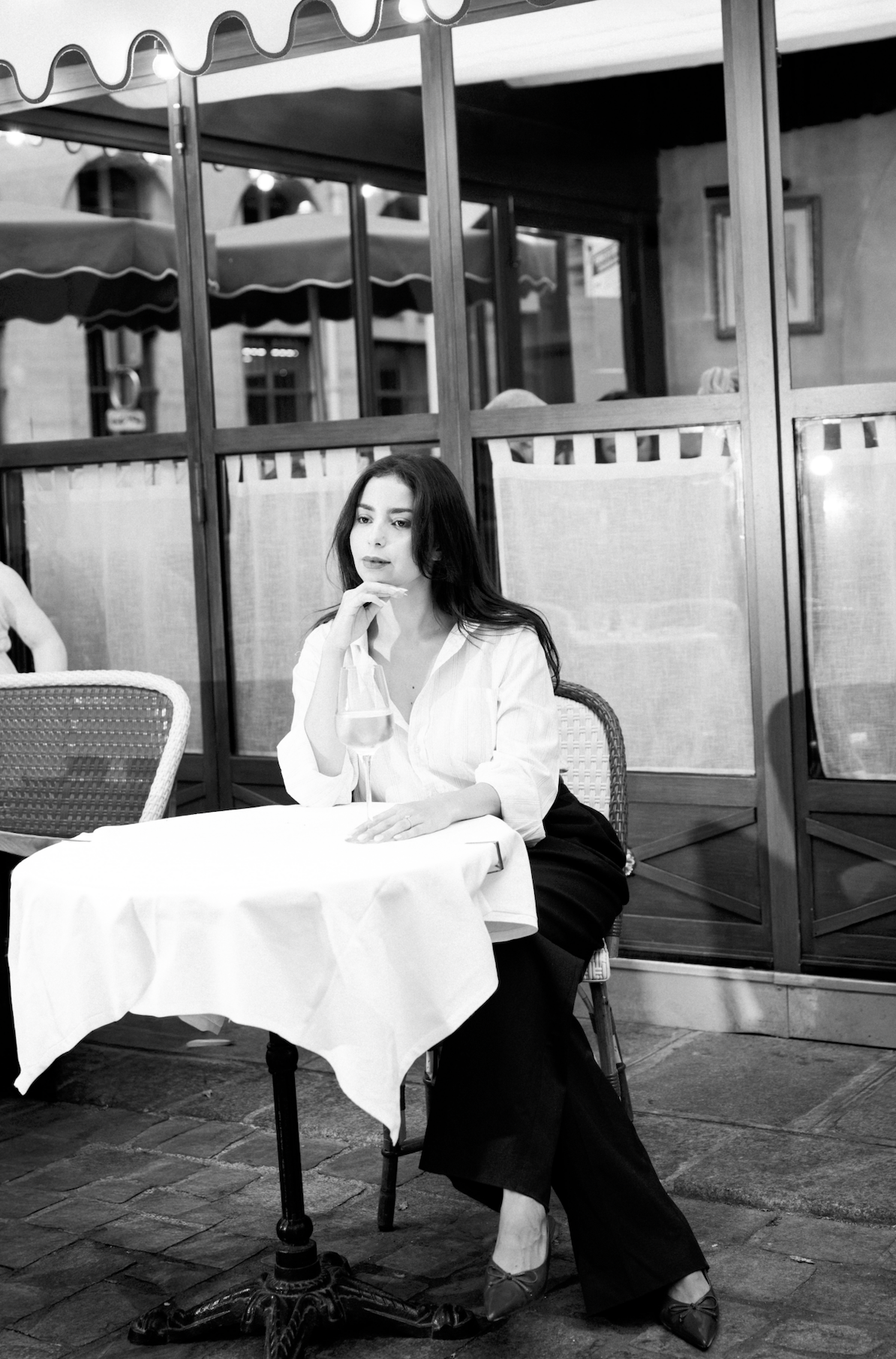 Une femme assise à une table dans un restaurant ou un café, tenant un verre de vin, en train de penser ou de regarder pensivement. L'ambiance est élégante, avec une décoration intérieure et une ambiance sophistiquée.