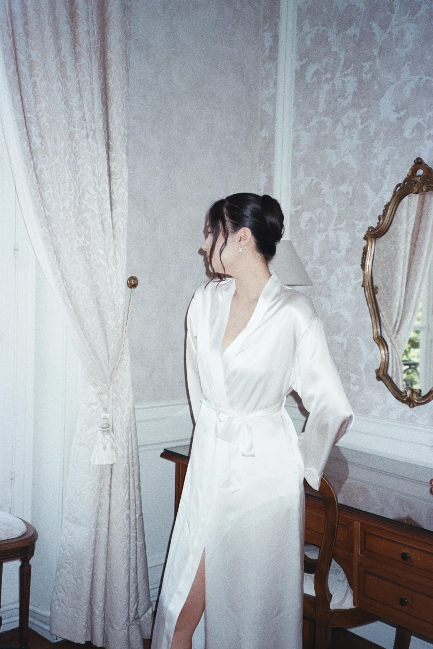 Une femme future mariée en peignoir blanc regarde de côté dans une pièce élégante avec mur en papier peint floral, miroir doré, et une table en bois.