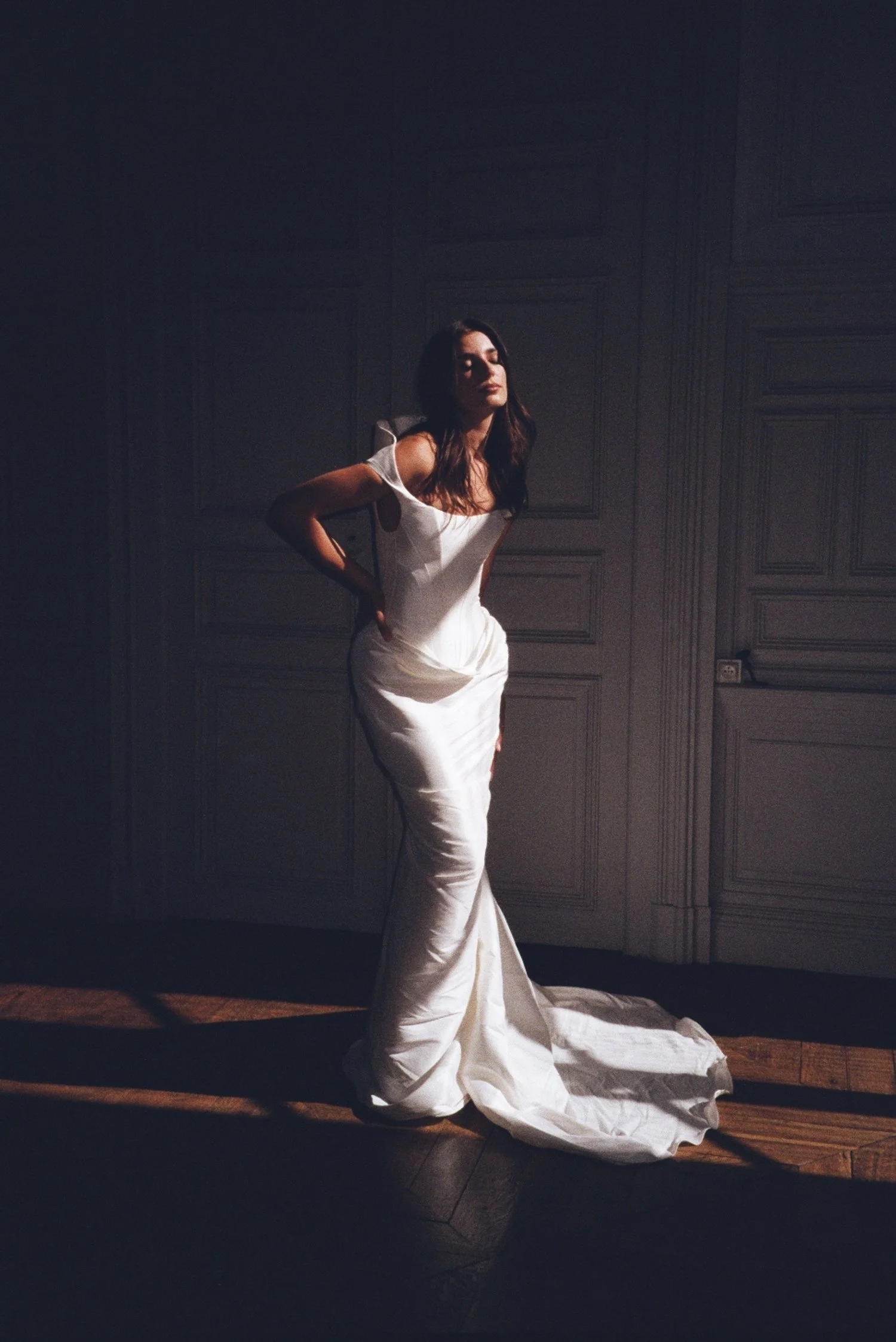 Une femme en robe blanche longue posant dans une pièce sombre avec des murs en bois, avec une lumière concentrée sur elle, créant un contraste avec l'obscurité environnante. Une mariée élégante et sophistiquée