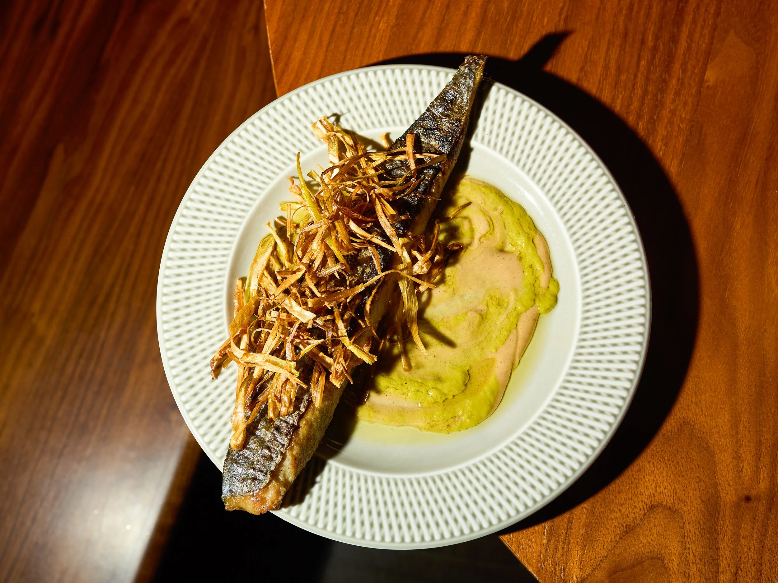 Grilled whole fish topped with crispy fried onions, served with green sauce on a white plate