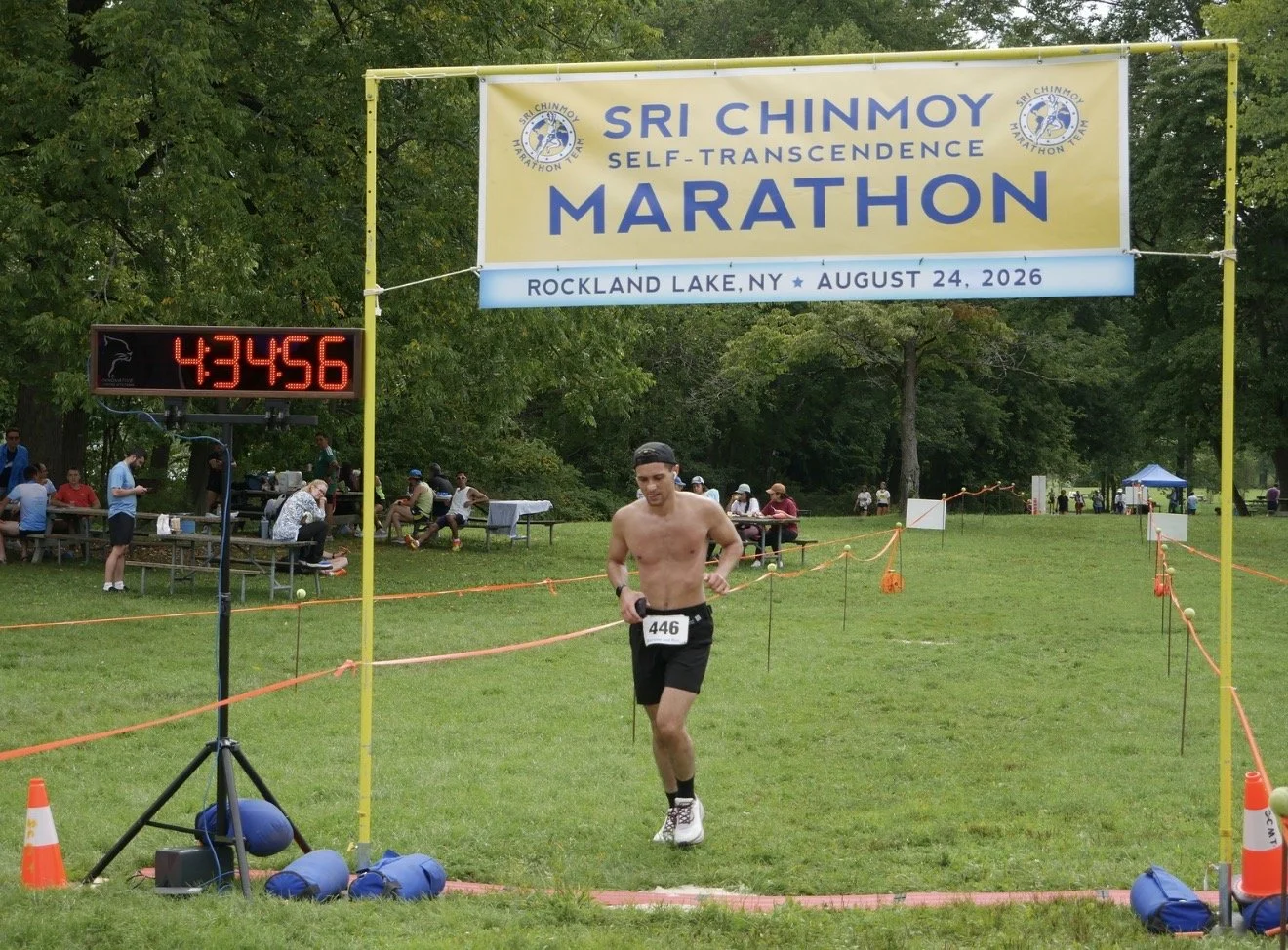 A male marathon runner wearing black shorts, white shoes, and a black cap crossing the finish line at the Sri Chinmoy Self-Transcendence Marathon held at Rockland Lake, NY, on August 24, 2026. The digital clock shows 4:34:56.