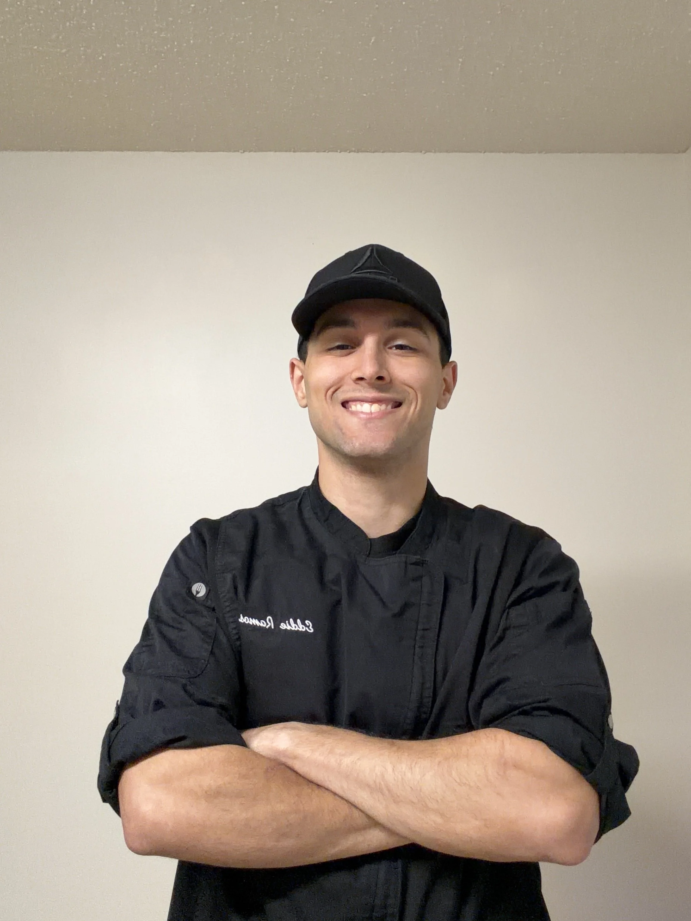 A smiling man in a black chef's jacket and black cap standing with his arms crossed in front of a plain white wall.