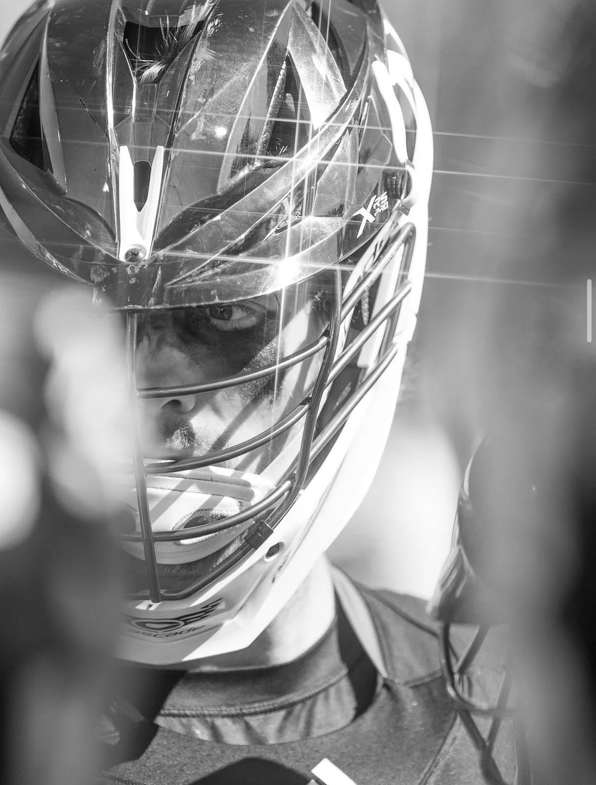 A black and white photograph of a lacrosse player wearing a helmet with a full face cage, partially covering his face. The player's eyes are visible through the cage, and he appears focused. The helmet has the brand logo 'X' and 'XS' on the side.