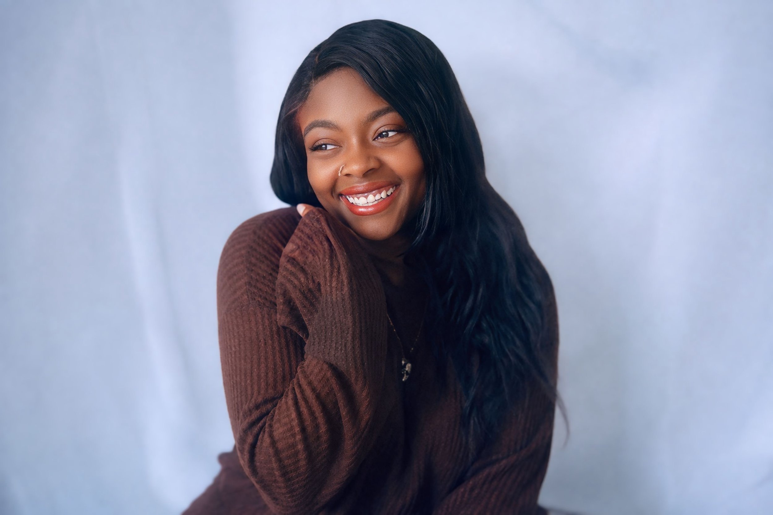 A woman with long black hair, smiling and wearing a brown sweater, posing against a light blue background.