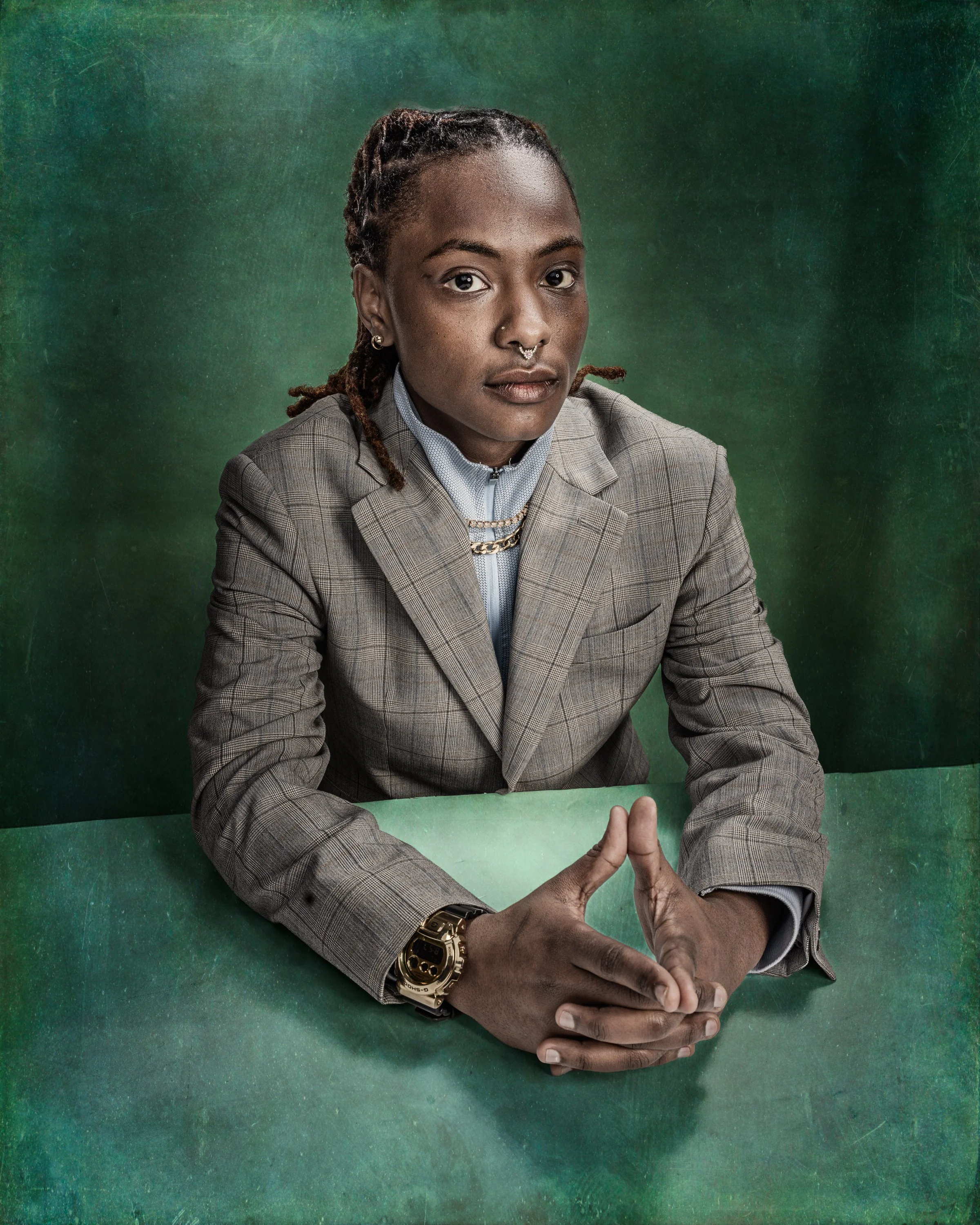 A portrait of a woman wearing a checked suit with a white shirt, with dreadlocked hair, gold jewelry, and a nose ring, sitting at a green table with a dark green background. Her hands are clasped in front of them on the table.